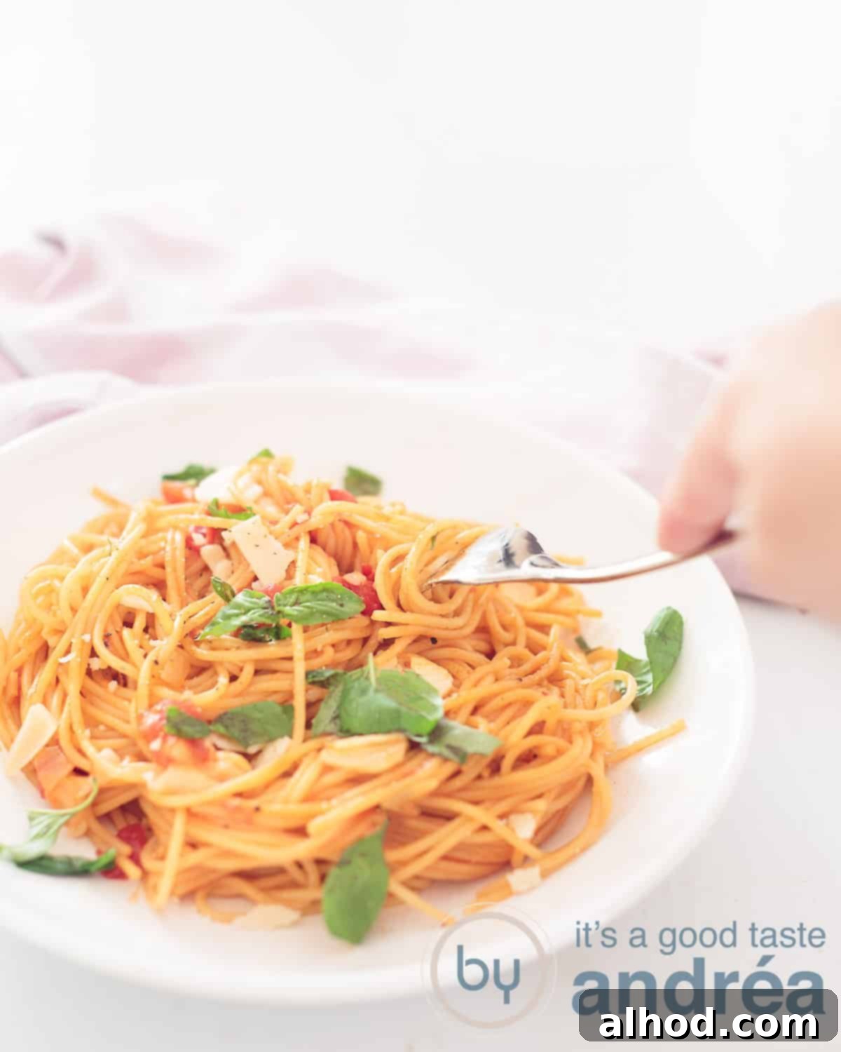 One Pan Veggie Pasta 2 An elevation photo with a white plate filled with pasta, cheese, tomato and basil at the bottom left. A fork pricks a bite. a white background. In the back left a pink and white tea deck