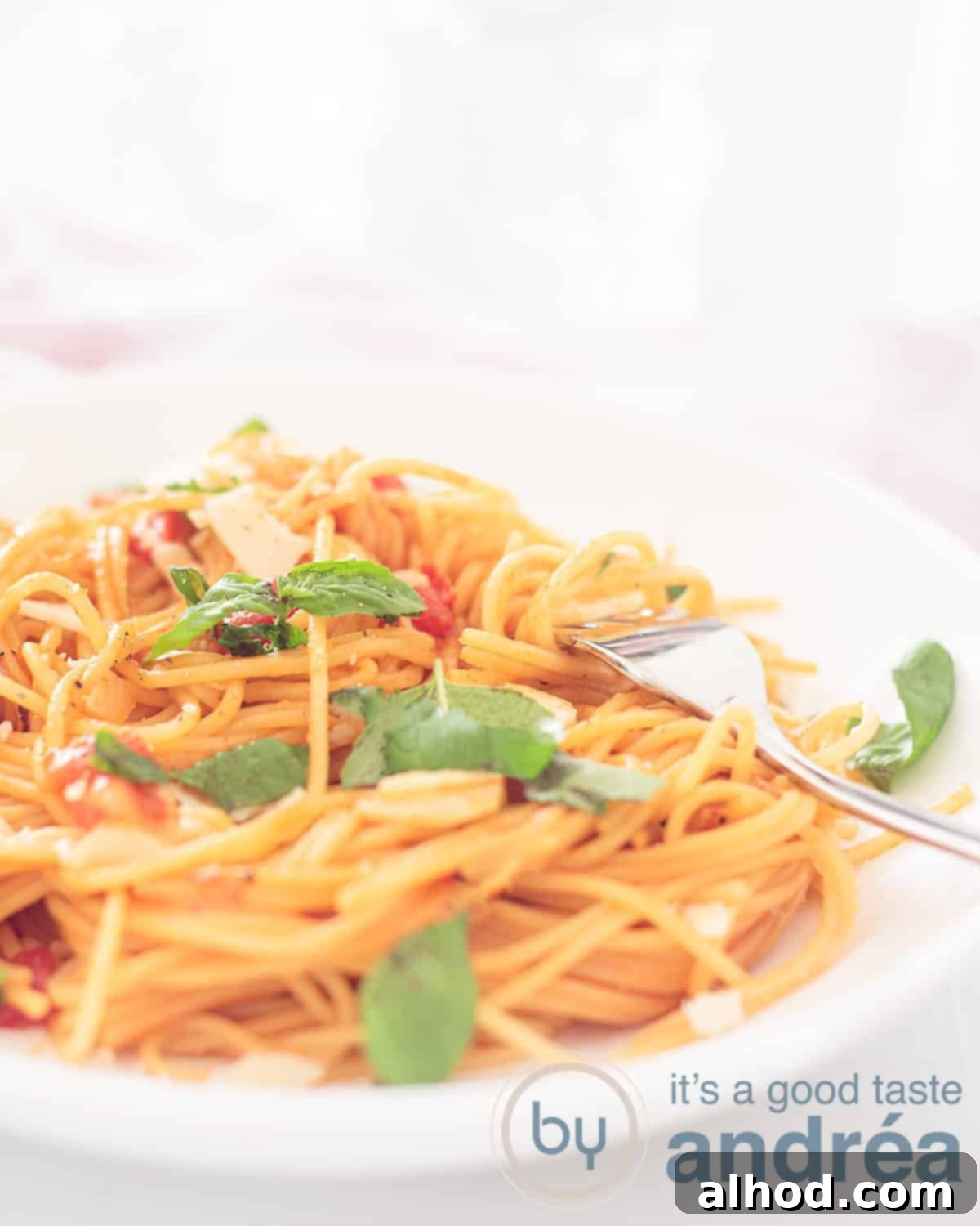 One Pan Veggie Pasta 5 An elevation photo showing part of a deep white plate with spaghetti, basil, cheese, and tomatoes. A gold fork pierces it. A white background.