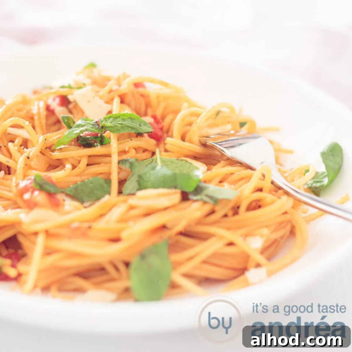 A square photo with a white plate filled with spaghetti, cheese, tomato and sprinkled with basil leaves. A gold fork sticks into it.