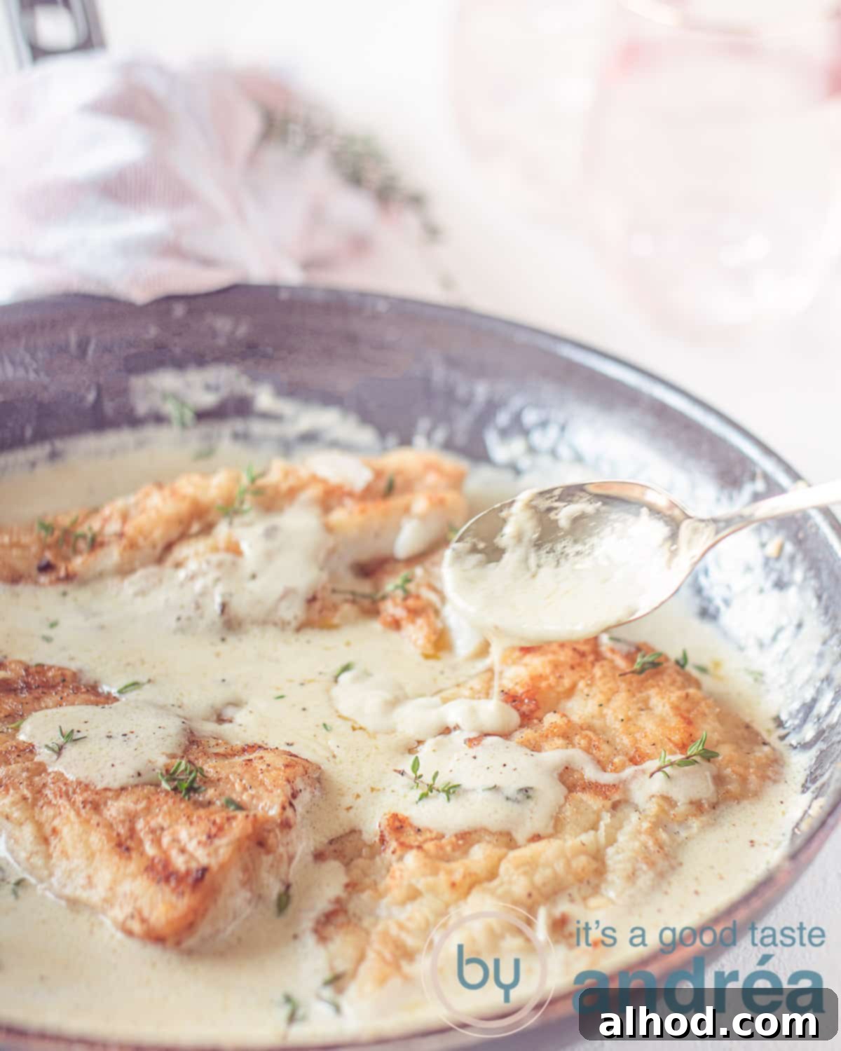 Cod Vin Blanc 5 An elevation view of a frying pan with golden-brown cod fillets being generously drizzled with white wine sauce, against a clean white background.