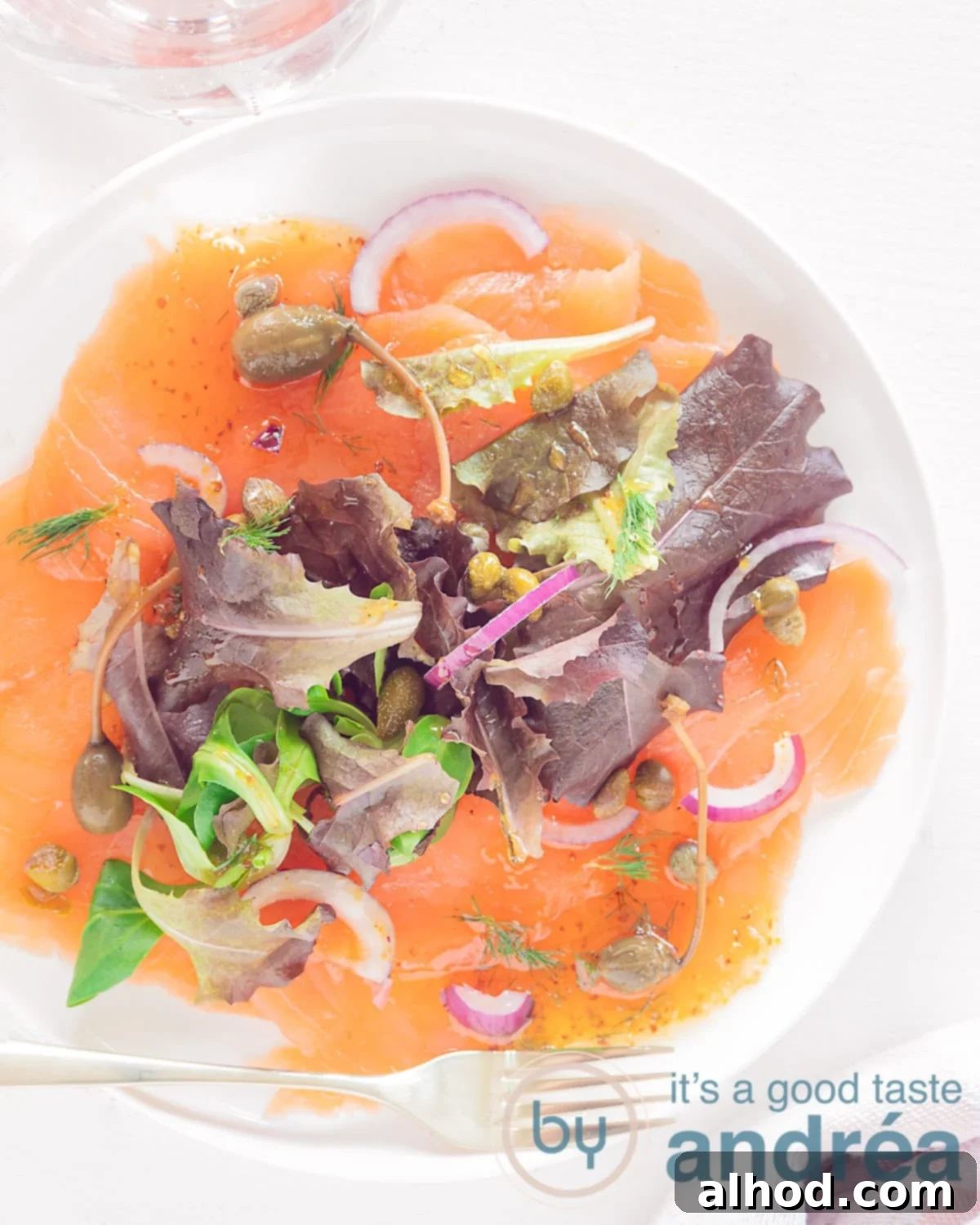 Silken Smoked Salmon Carpaccio 5 A photo from above of a round white plate with slices of smoked salmon carpaccio, salad, red onion, capers and a honey dill sauce. A white background.