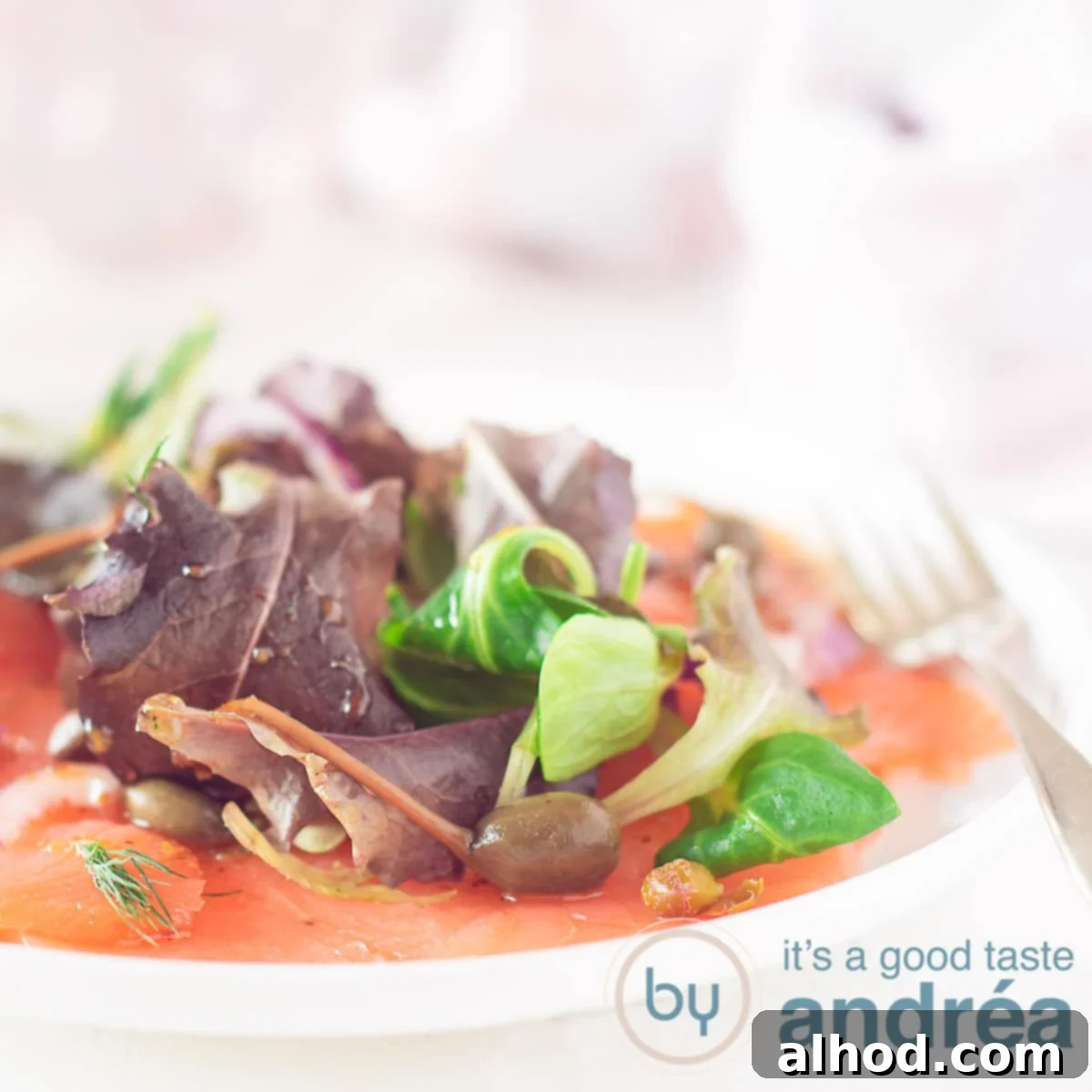 Silken Smoked Salmon Carpaccio 6 A square photo with part of a plate covered with salmon carpaccio, lettuce, red onion, capers and honey mustard dressing. A white background.