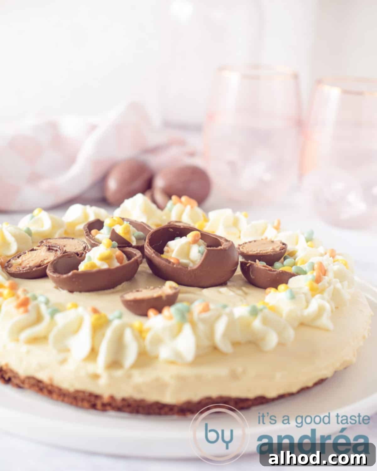 A high-angle photo of an Easter cake with eggnog, decorated with pastel Easter eggs and dollops of whipped cream, on a white background with chocolate eggs scattered around.