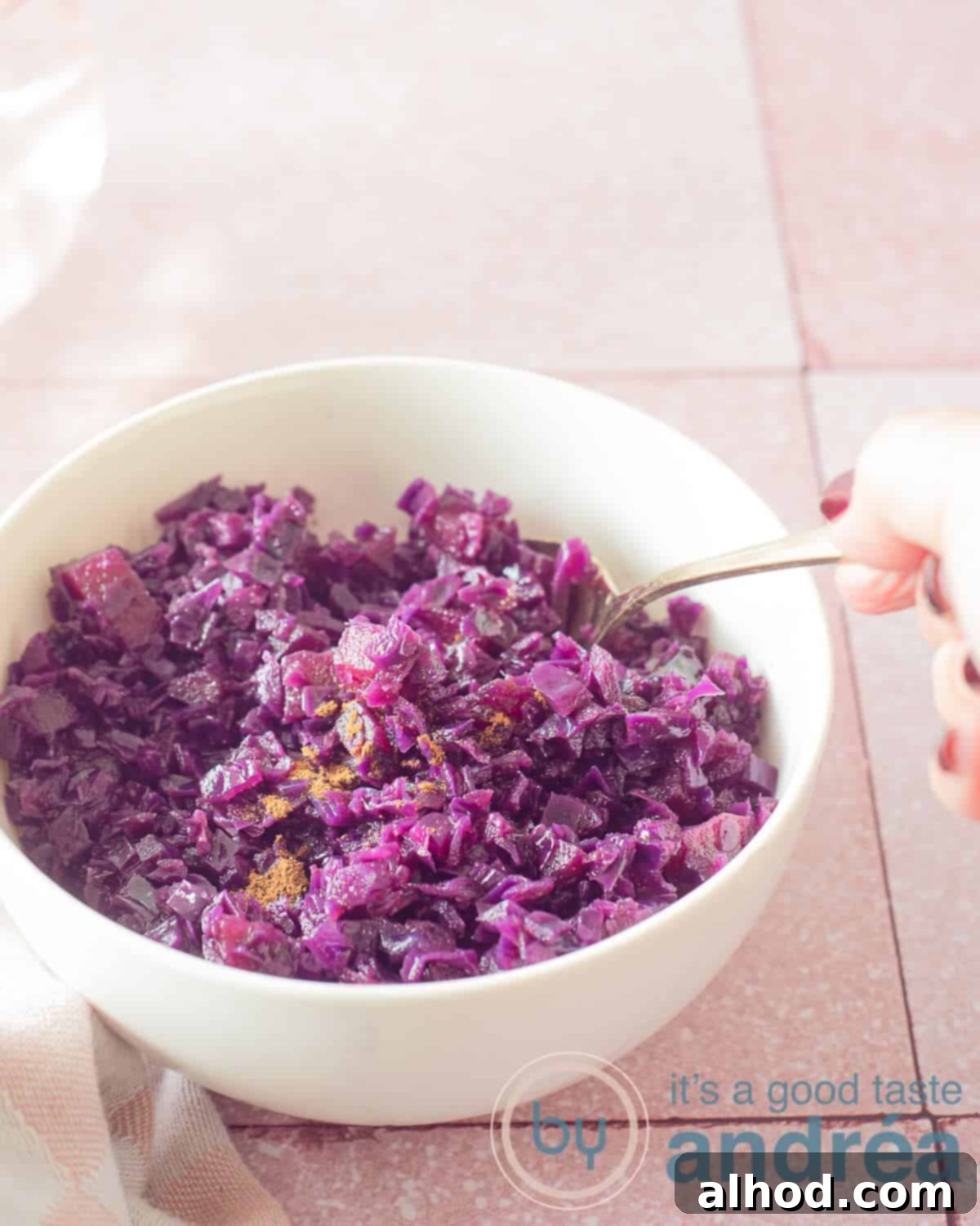 Ruby Red Balsamic Apple Braise 2 An elevation photo showing part of a white bowl of red cabbage. A golden spoon scoops cabbage out of the bowl. A pink background.