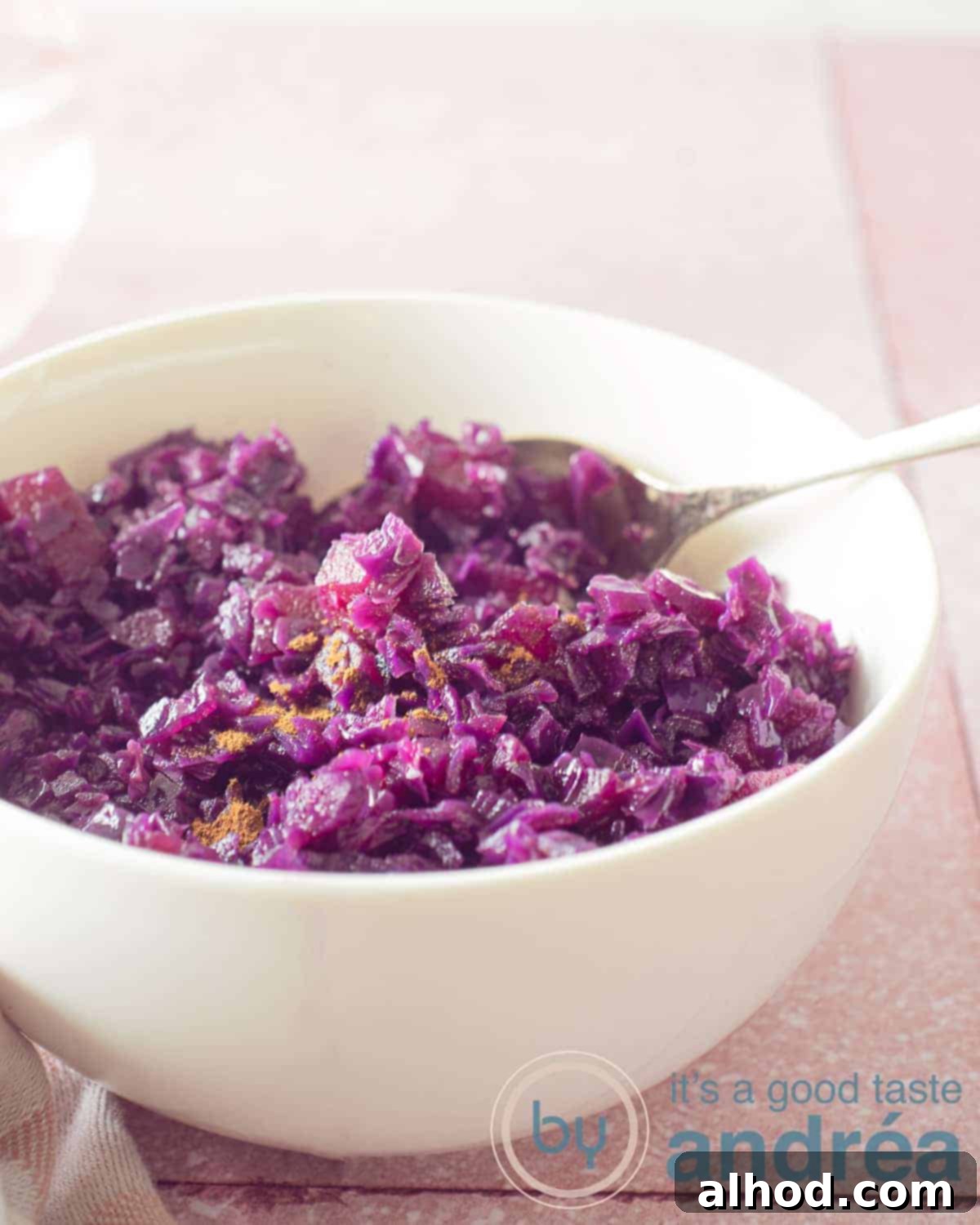 Ruby Red Balsamic Apple Braise 5 A height photo with a pink background, on top of which is a white bowl filled with red cabbage with an apple.