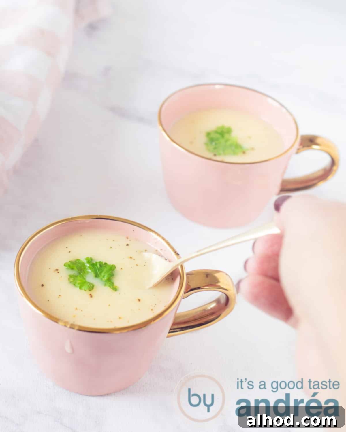 Velvety Asparagus Soup 2 A height shot of two pink cups filled with white asparagus soup, garnished with parsley. A golden spoon takes a bite. A marbled white background.