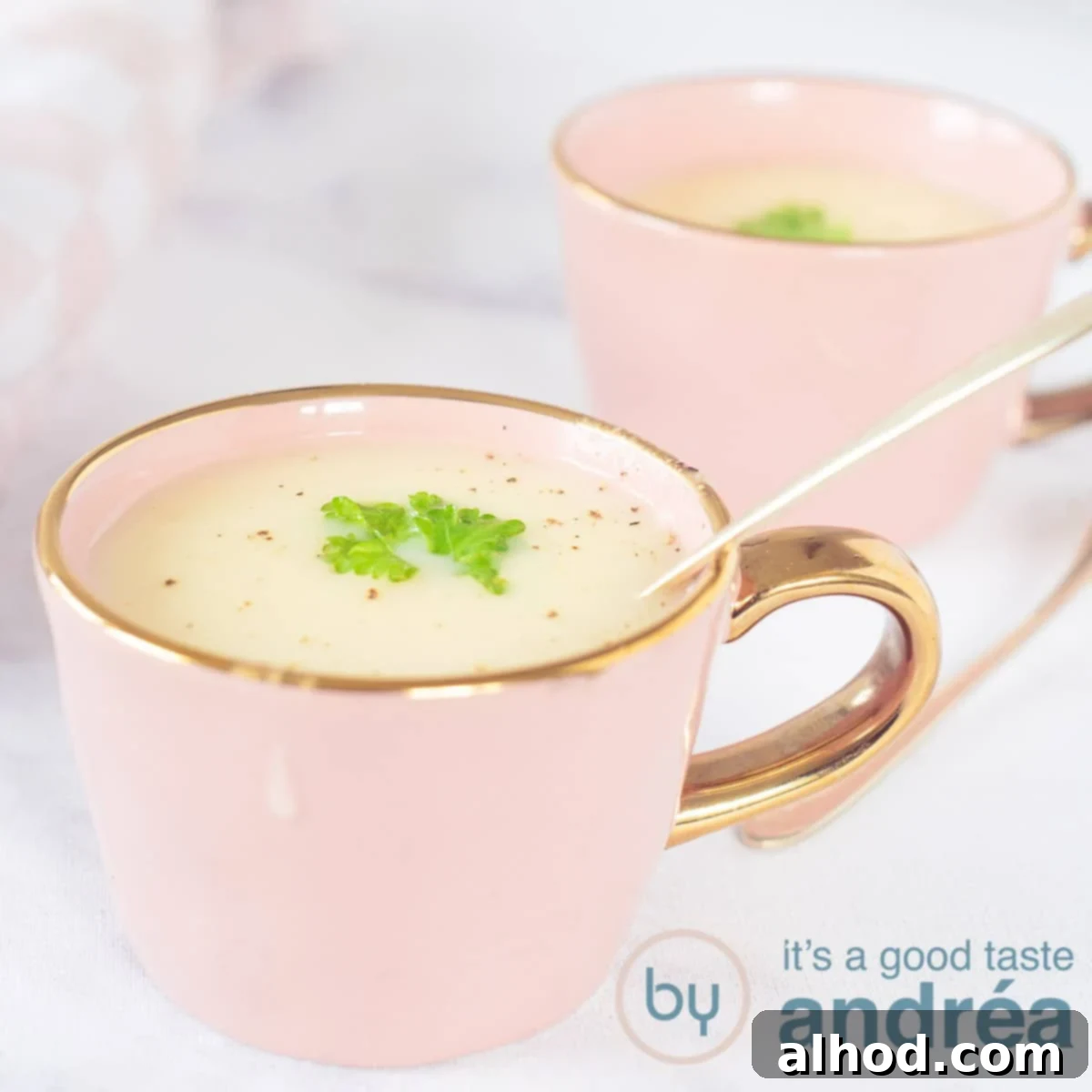 Velvety Asparagus Soup 6 A square photo, two pink cups diagonally behind each other. In it a creamy asparagus soup with parsley. A white marbled background.