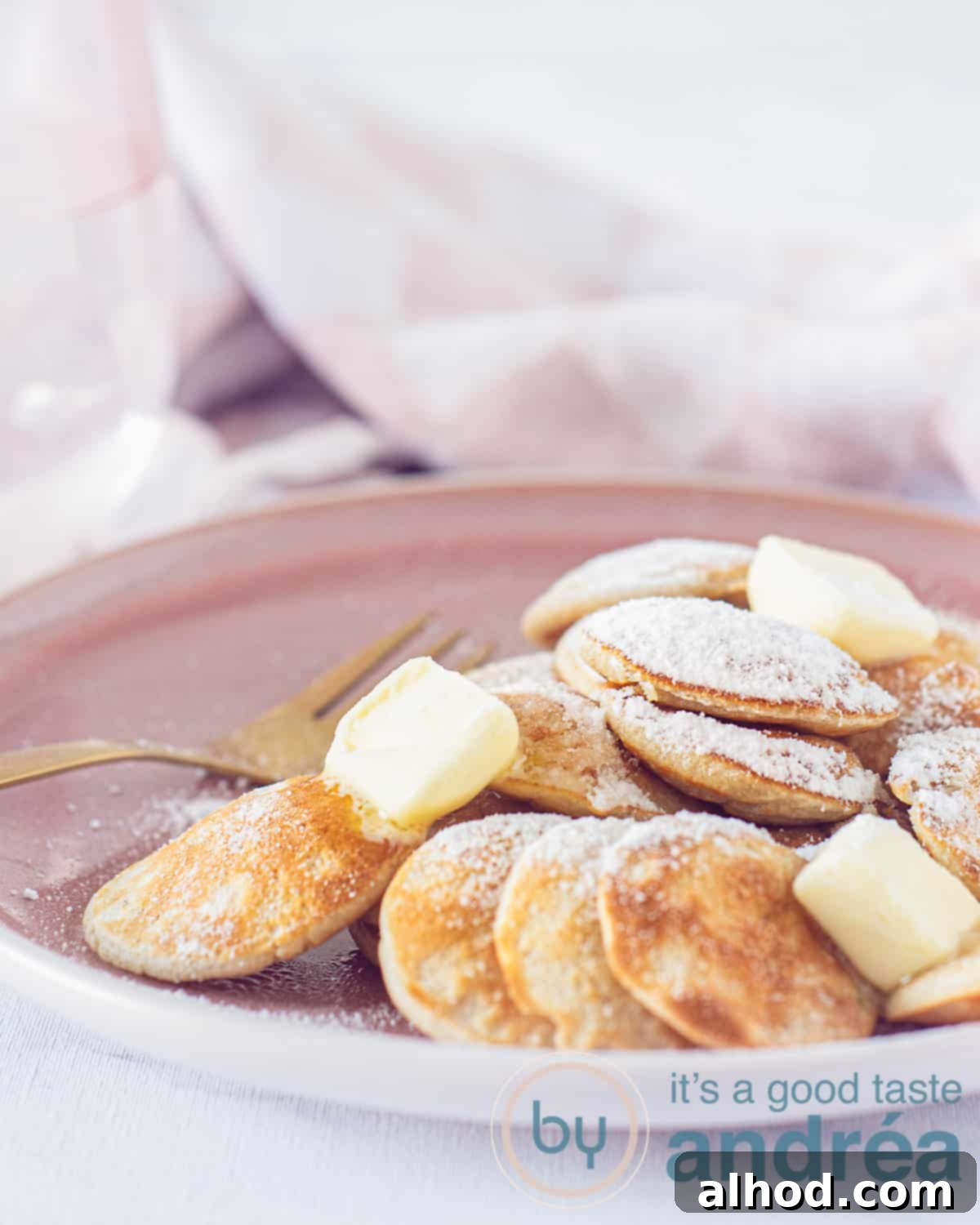 Poffertjes Dutch Mini Pancakes 2 An elevation photo with part of a pink plate with a pile of poffertjes, butter and icing sugar. In the background a pink and white tea towel.