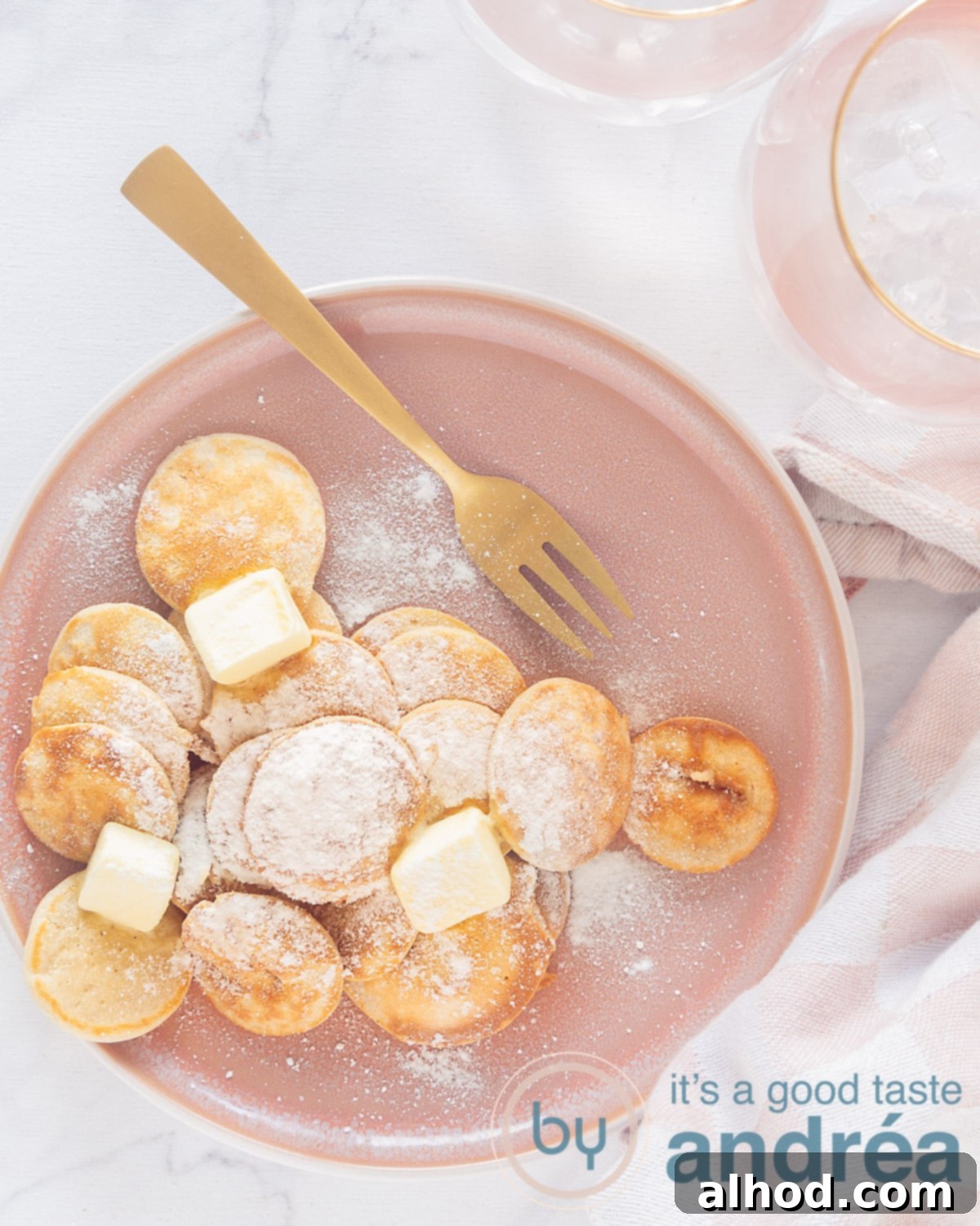Poffertjes Dutch Mini Pancakes 5 A photo from above, in height, of a pink plate with poffertjes, icing sugar and butter. A golden fork lies above it. A white background. At the top right part of two pink glasses.