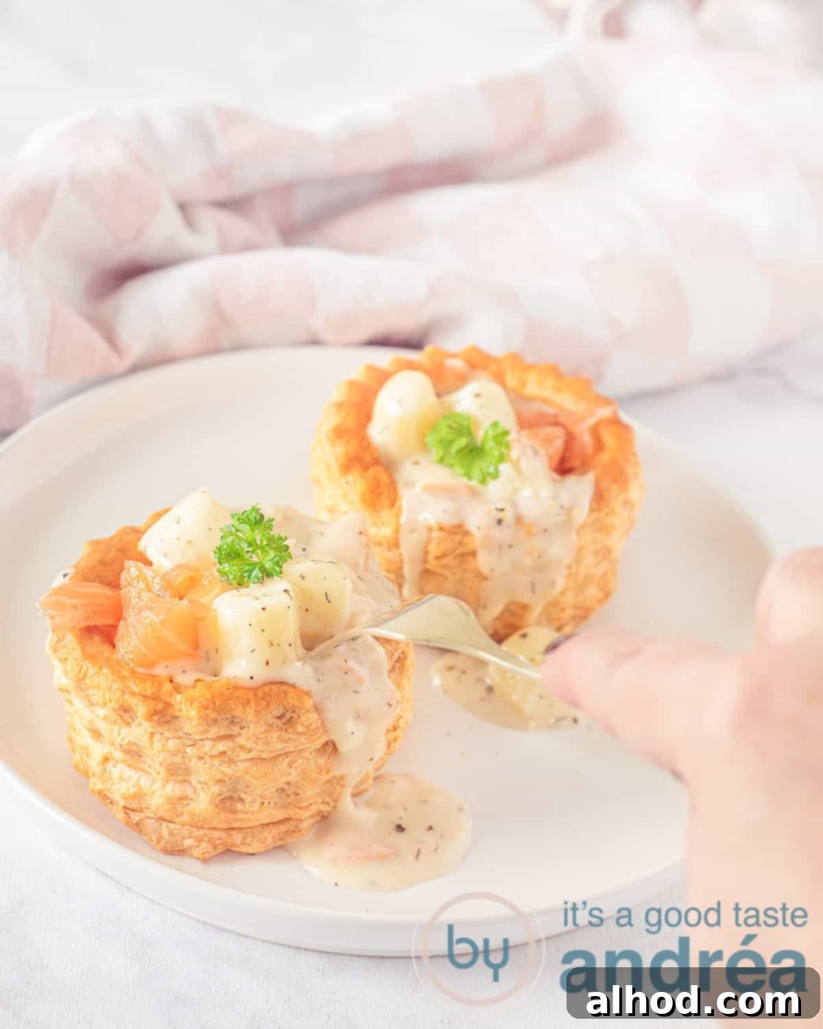 An elevation photo with a white plate with two ragout trays with asparagus ragout and salmon. A fork pierces the ragout. In the background a pink and white checkered tea towel.