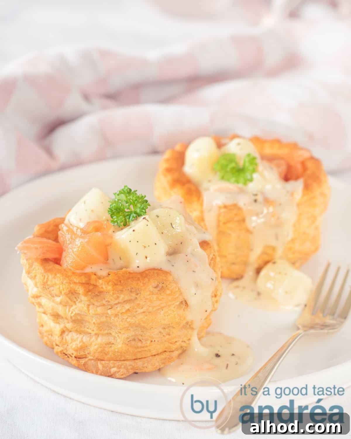 Flaky Pastry Asparagus Ragout 5 An elevation photo of two puff pastry cases filled with asparagus with smoked salmon ragout on a white plate. A golden fork lies beside it. In the background a white pink checkered tea towel