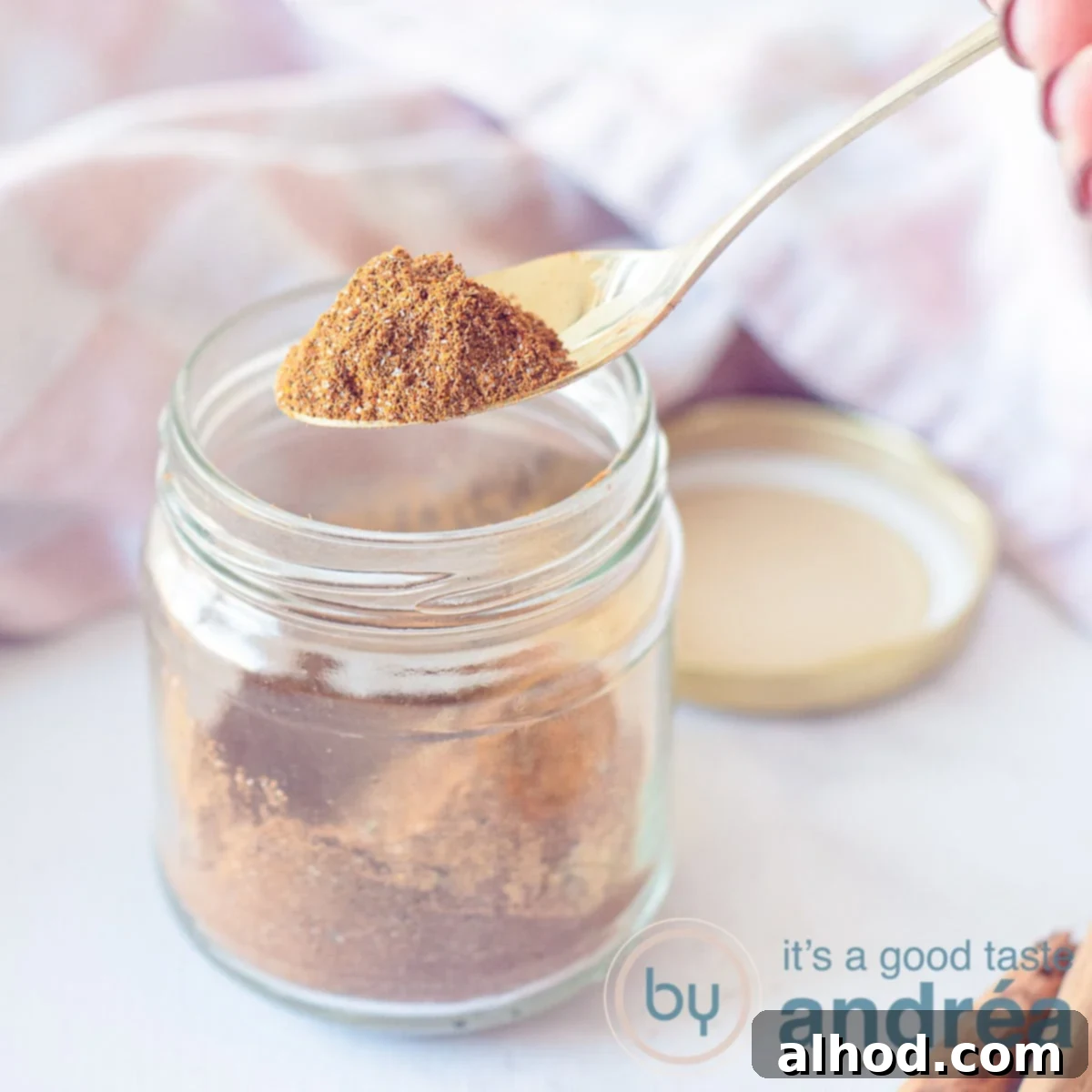 Homemade Shawarma Spice Mix 5 A square photo with a glass jar filled with shawarma spices. A pink white tea towel in the background. A spoon scoops the mix out of the jar, ready for use.