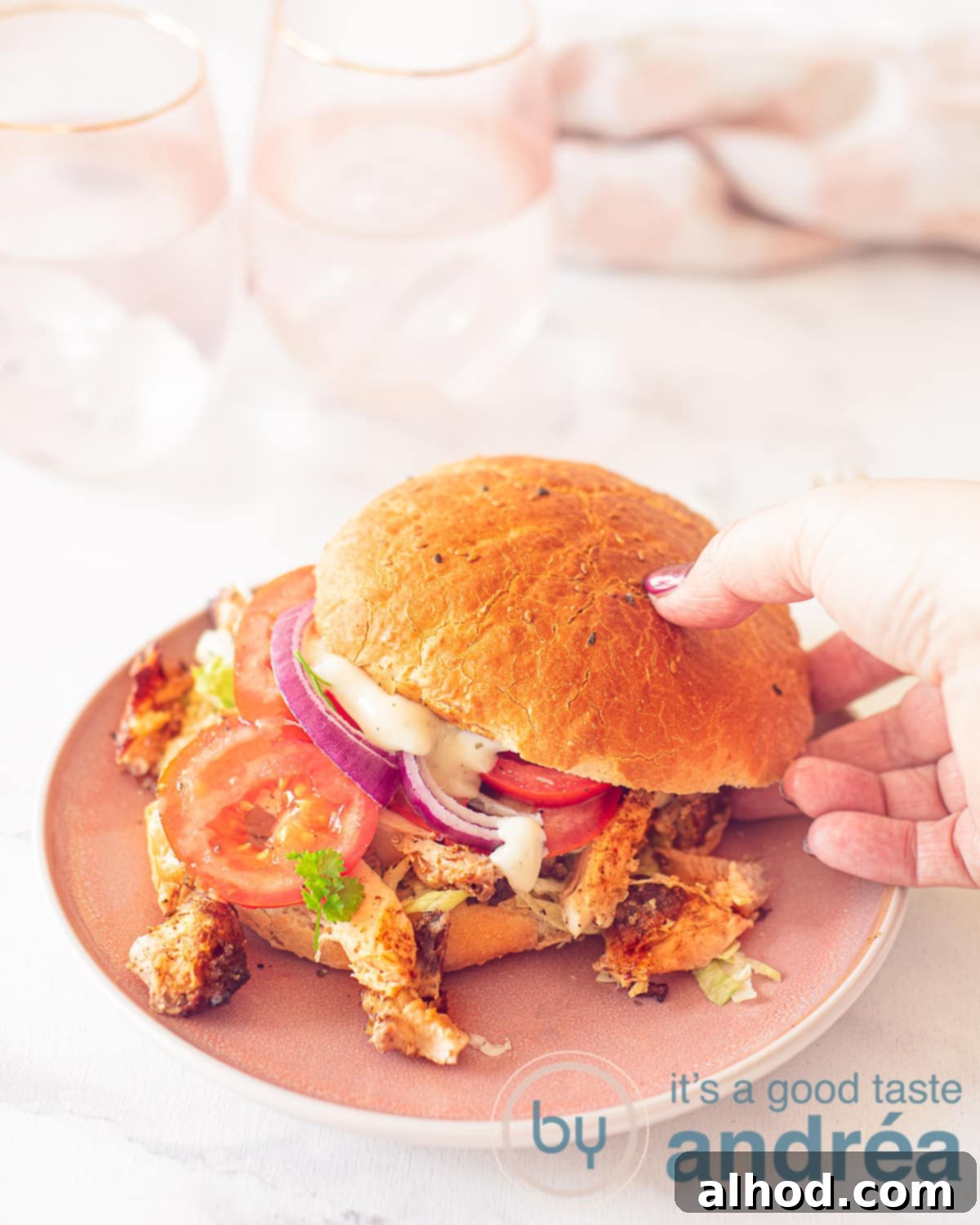 A height photo of a chicken kebab sandwich, raw vegetables on a pink plate. A hand takes the sandwich. A white surface in the background a pink white tea towel
