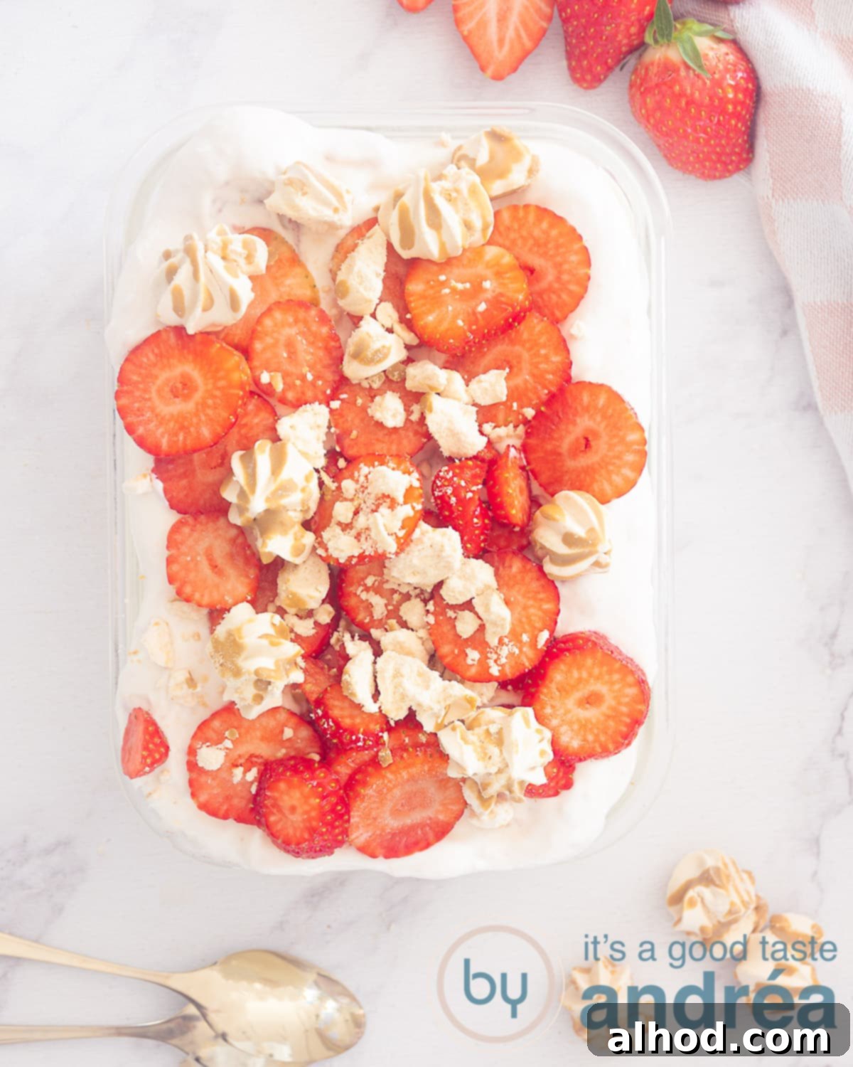 Luscious Egg-Free Strawberry Tiramisu 2 A photo from above of a bowl of strawberry tiramisu topped with strawberry slices and crumbled meringue. At the bottom two golden spoons and meringue. Extra strawberries on top. A white background.