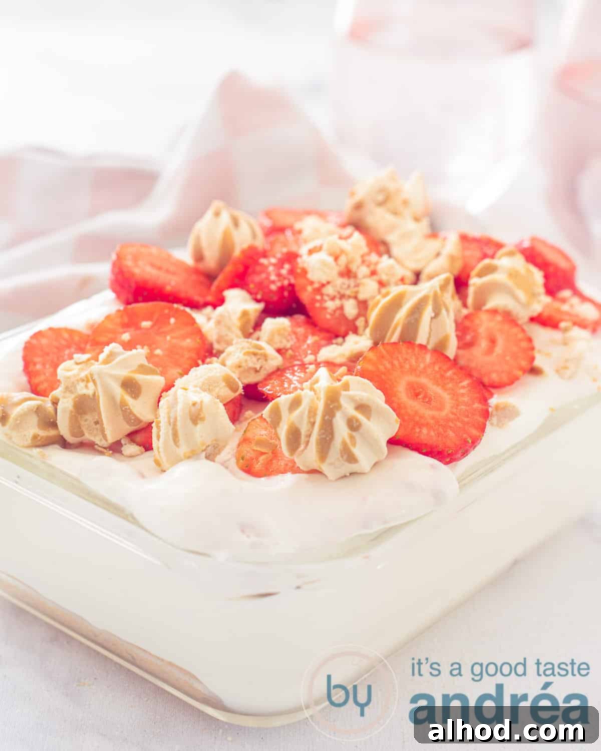 Luscious Egg-Free Strawberry Tiramisu 5 An elevation view of a glass bowl filled with strawberry tiramisu with a strawberry and meringue garnish. a white background and a pink and white tea towel in the background
