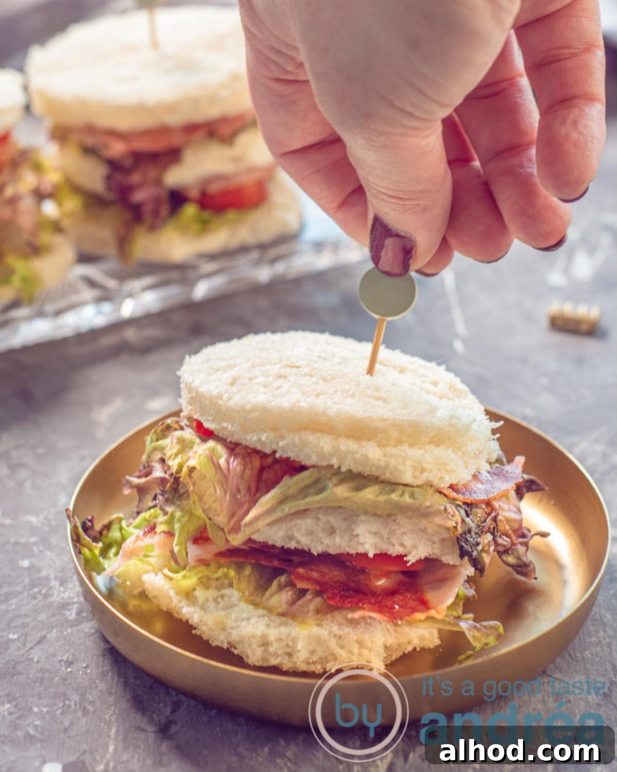 BLT Club Bites 5 A photo in height with a gold plate with a BLT mini Sandwich on it. A hand sticks a tapas picker in it. More sandwiches in the background