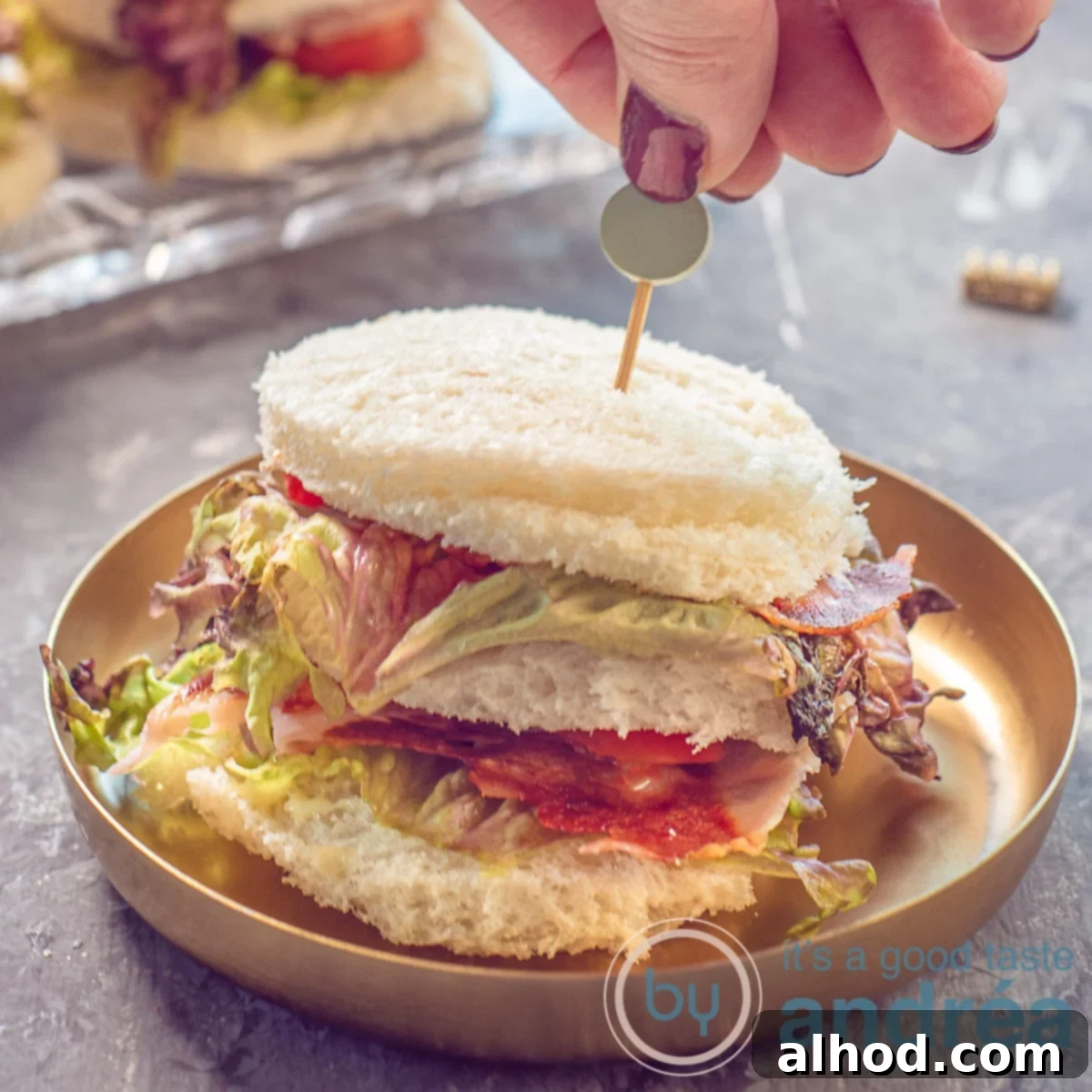 BLT Club Bites 6 A square photo with a BLT mini sandwich on a gold plate on a gray background. A hand sticks a skewer in it.