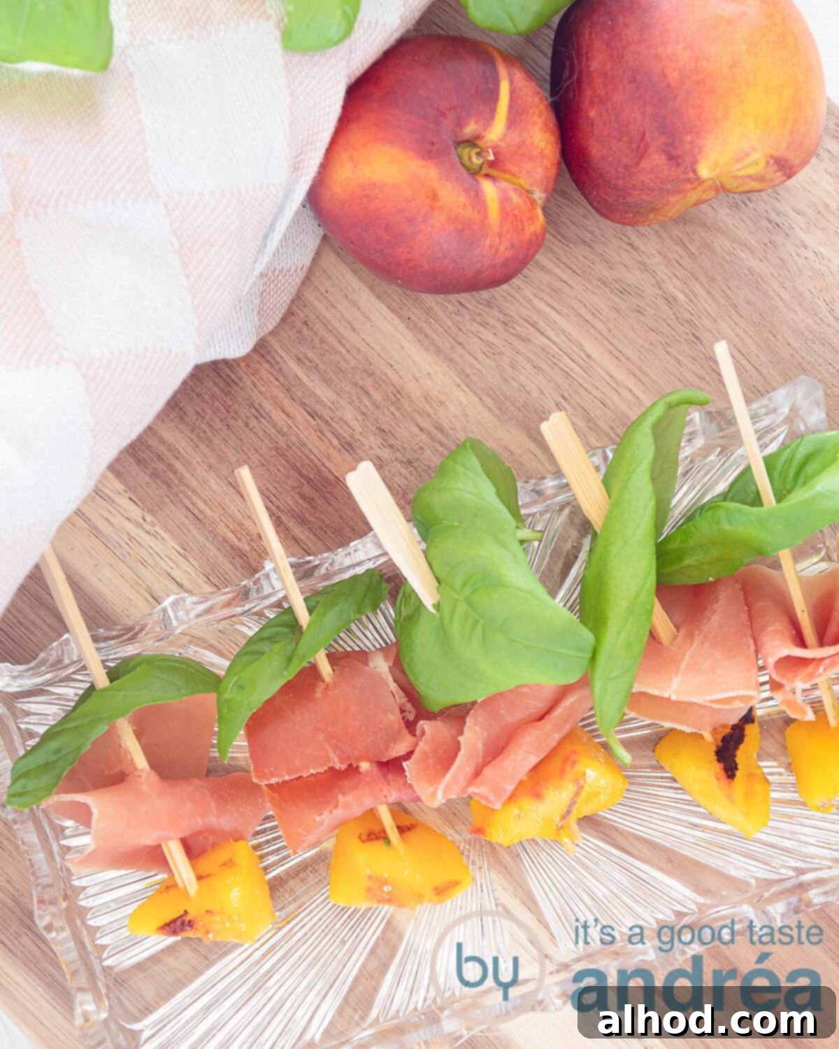 Serrano Ham Wrapped Grilled Peaches 3 A top-down view of grilled peach with ham and basil arranged on a glass dish, set on a rustic wooden board. Two fresh peaches and a pink and white tea towel complete the aesthetic in the background.
