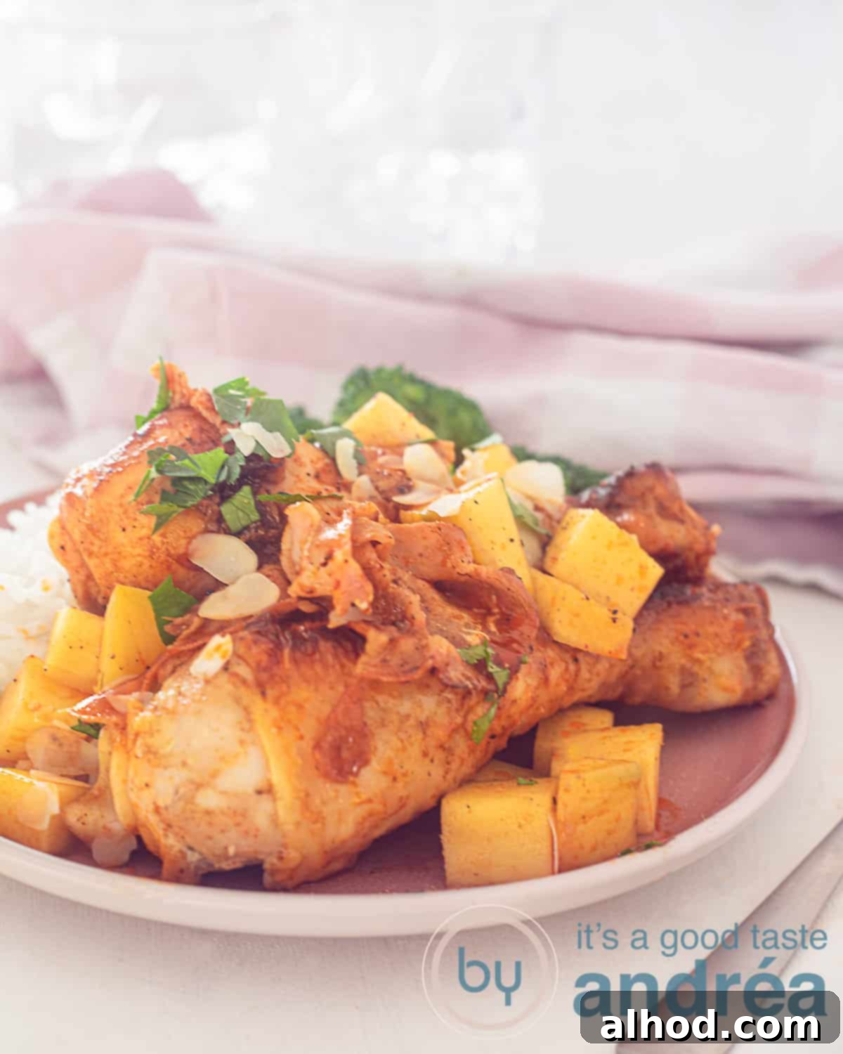 Mango-Glazed Chicken Drumsticks with Bacon 2 An elevation photo with a pink plate with drumsticks, mango cubes in sauce and broccoli. A pink white tea towel in the background.