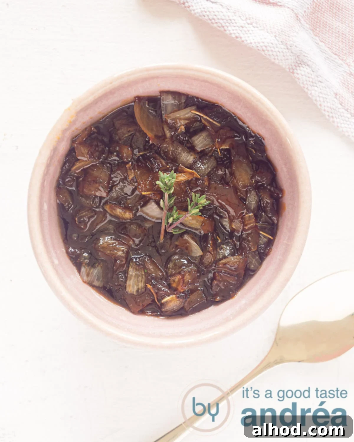 A photo from above of a pink bowl of caramelized onions garnished with fresh thyme on a white surface. At the bottom right a golden spoon.