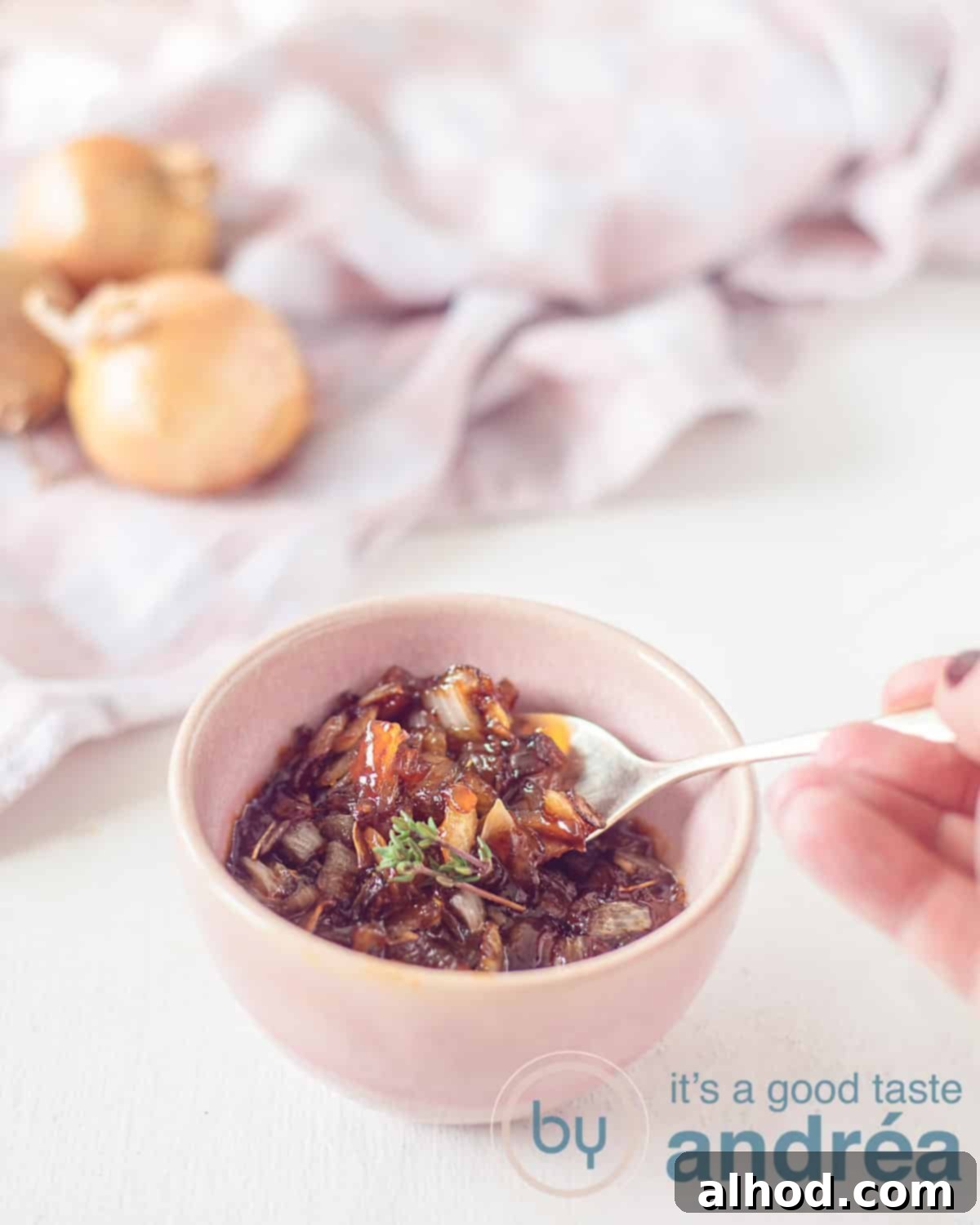 A white background with a pink dish filled with deliciously caramelized onions. In the background a pink and white tea towel and three onions.