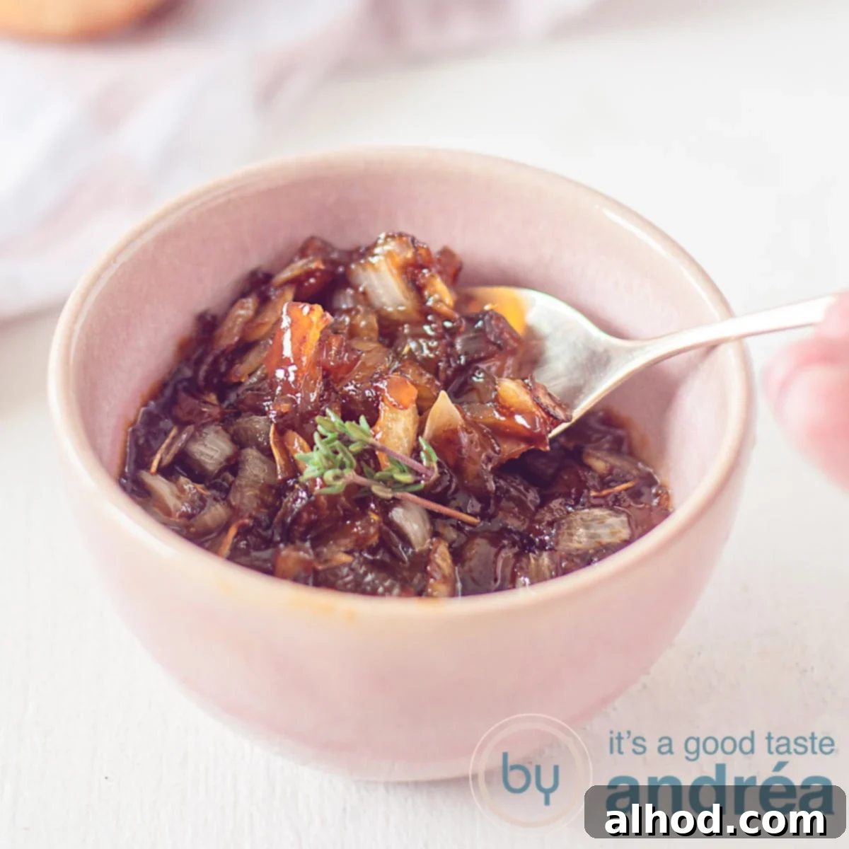 A square photo with a pink bowl filled with caramelized onion sauce. A spoon scoops some out. A white background