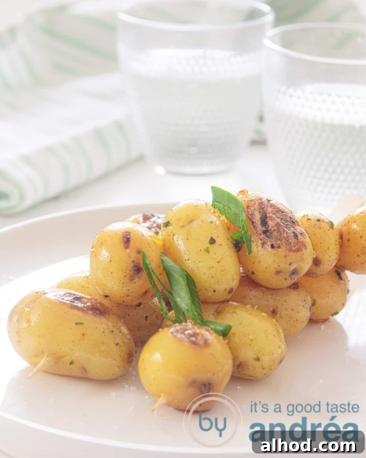 A height photo with a white plate with three skewers with delicious seasoned baby potatoes from the bbq. A white background. In the background two glasses of water and a white and green striped tea towel.