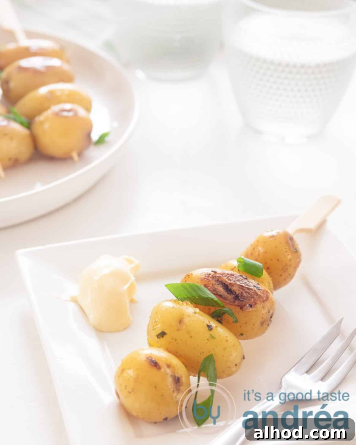 A baby kebab on a white plate with some sauce. In the background more baby potatoes on a stick and a glass of water.