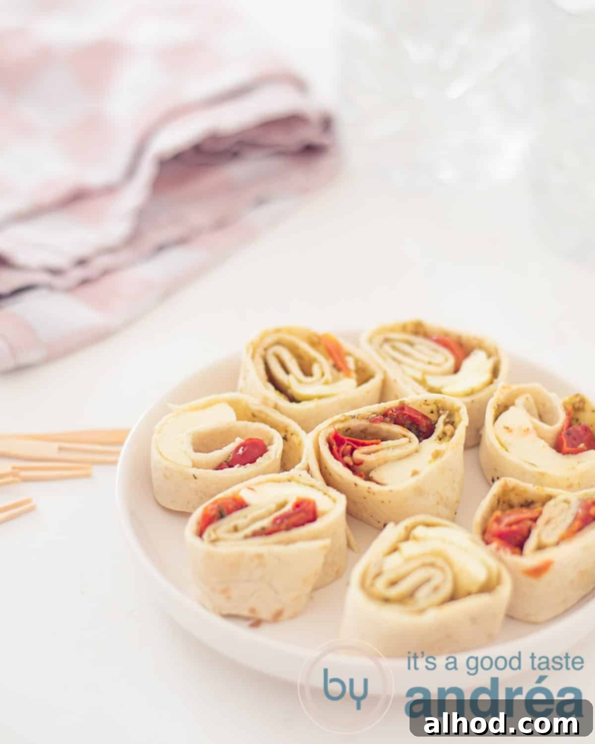 Caprese Swirls 5 An elevation photo with a white small round plate filled with Caprese wrap snacks at the bottom right. On the left some wooden tapas sticks. A white background. Top left a tea towel with pink and white.