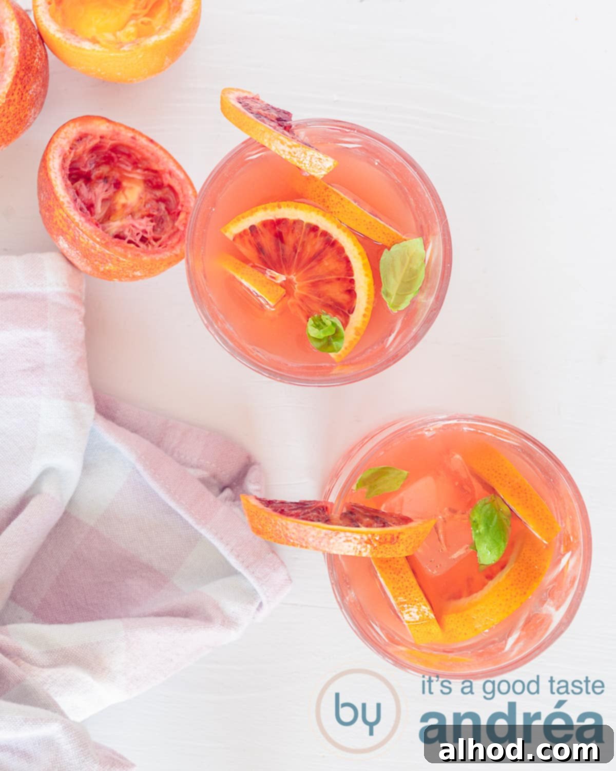 Crimson Citrus Chill 2 A photo from above of two glasses of iced tea with blood orange and mint. A white background. At the bottom left a pink white tea towel. Squeezed halves of blood orange at the top left.