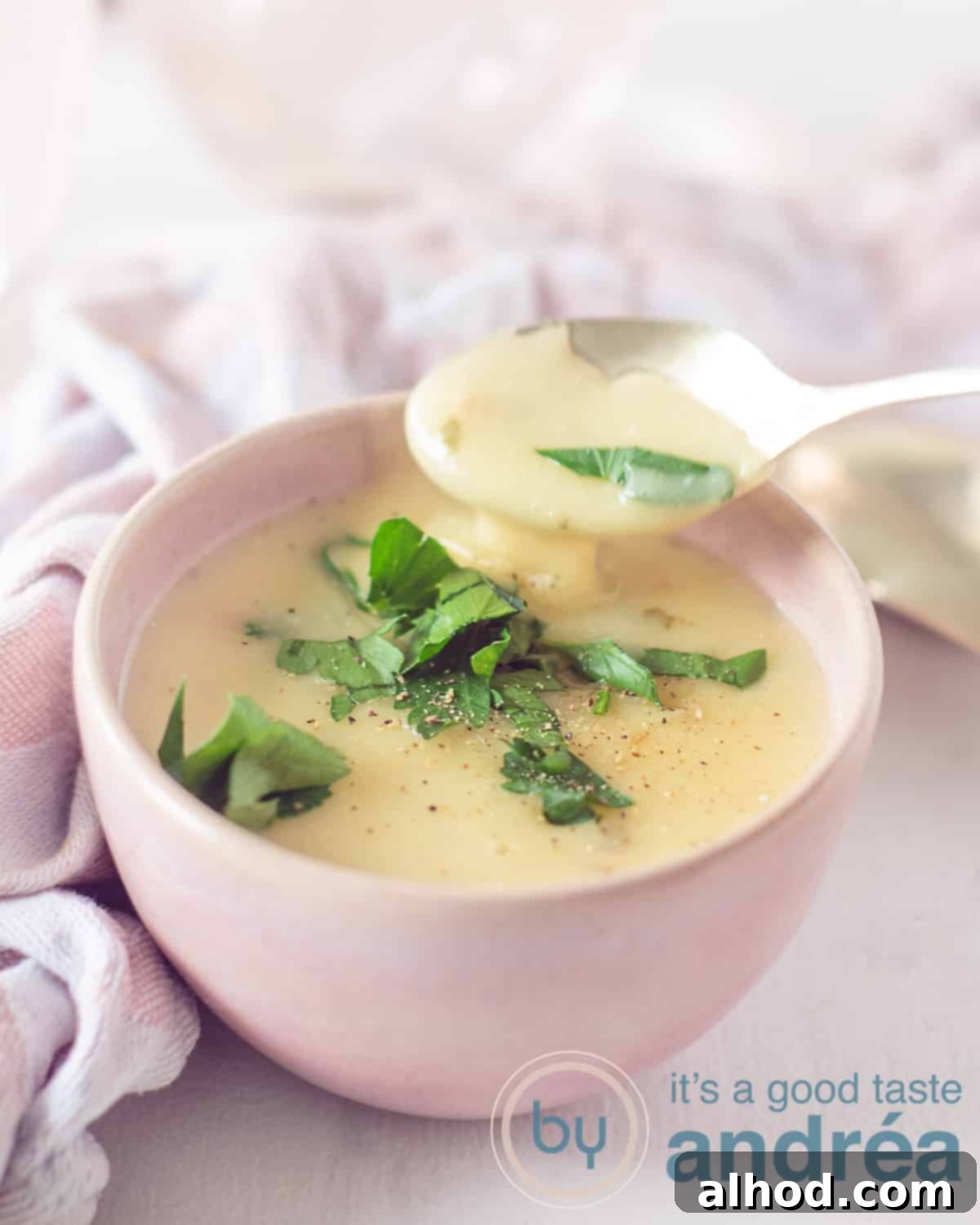 The Ultimate Garlic Parmesan Sauce 5 A height shot of a pink container filled with cheese sauce with Parmesan and garlic, garnished with parsley. A spoon scoops out sauce. A pink white tea towel in the background.