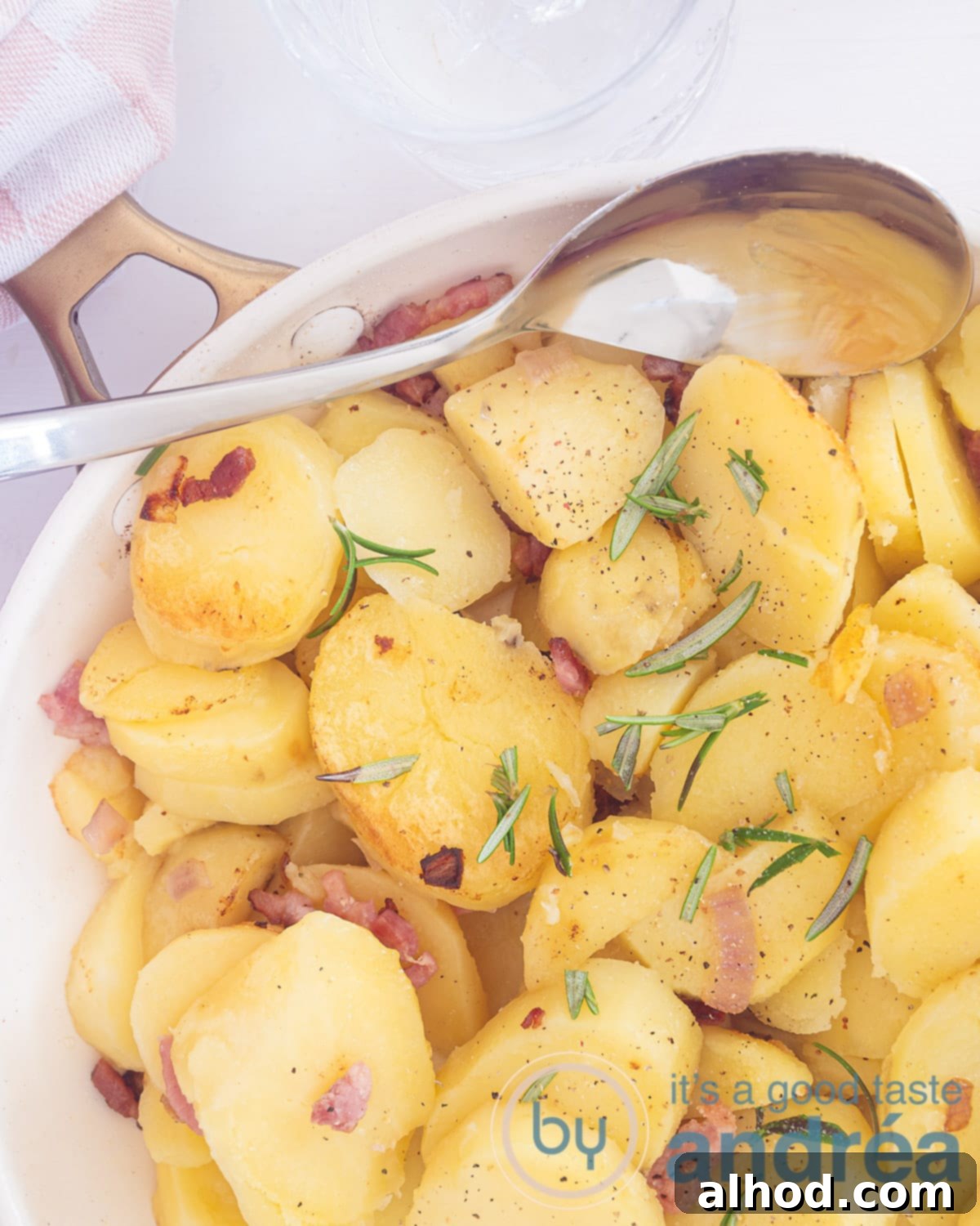 Classic German Fried Potatoes 2 A photo from above of a white frying pan with fried potatoes, onion, rosemary and bacon bits. A spoon scoops it in.