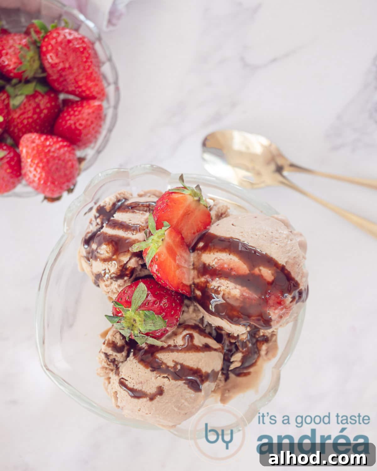Chocolate Strawberry Froyo Treat 2 A photo from above of a coupe filled with creamy chocolate Greek yogurt ice cream, generously topped with fresh strawberries and a drizzle of chocolate sauce. Top left, a glass dish with extra strawberries. Right, two golden spoons rest on a white background.