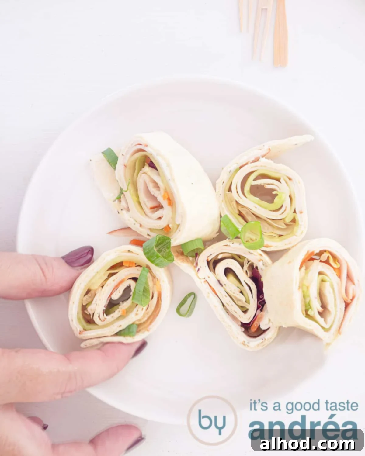 Creamy Chicken Tortilla Roll-Ups 2 A photo from above showing five wraps with chicken, salad and herb cheese. A hand takes a bite. A white background.