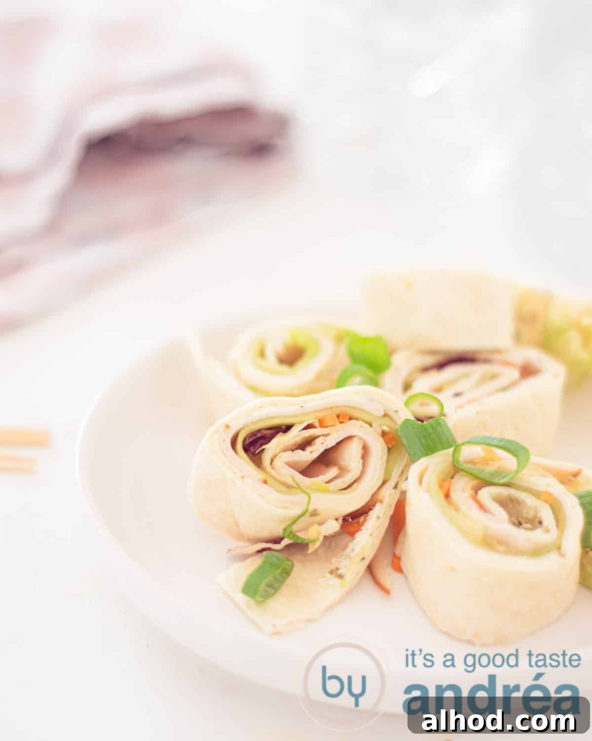 Creamy Chicken Tortilla Roll-Ups 5 A height photo with part of a white plate with five chicken wrap snacks with herb cheese and vegetables. A white background. In the back left a pink white tea towel.