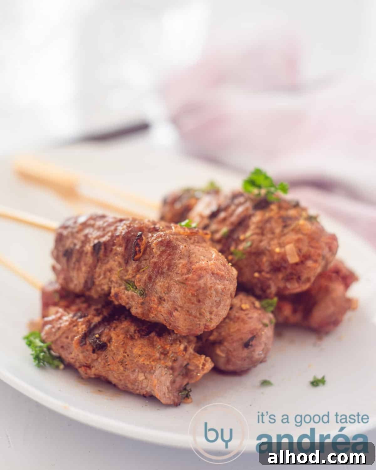 Authentic Moroccan Köfte Kebabs 2 A height photo with a white bowl with five ground beef skewers, deliciously seasoned on a white background, In the background a pink and white tea towel.