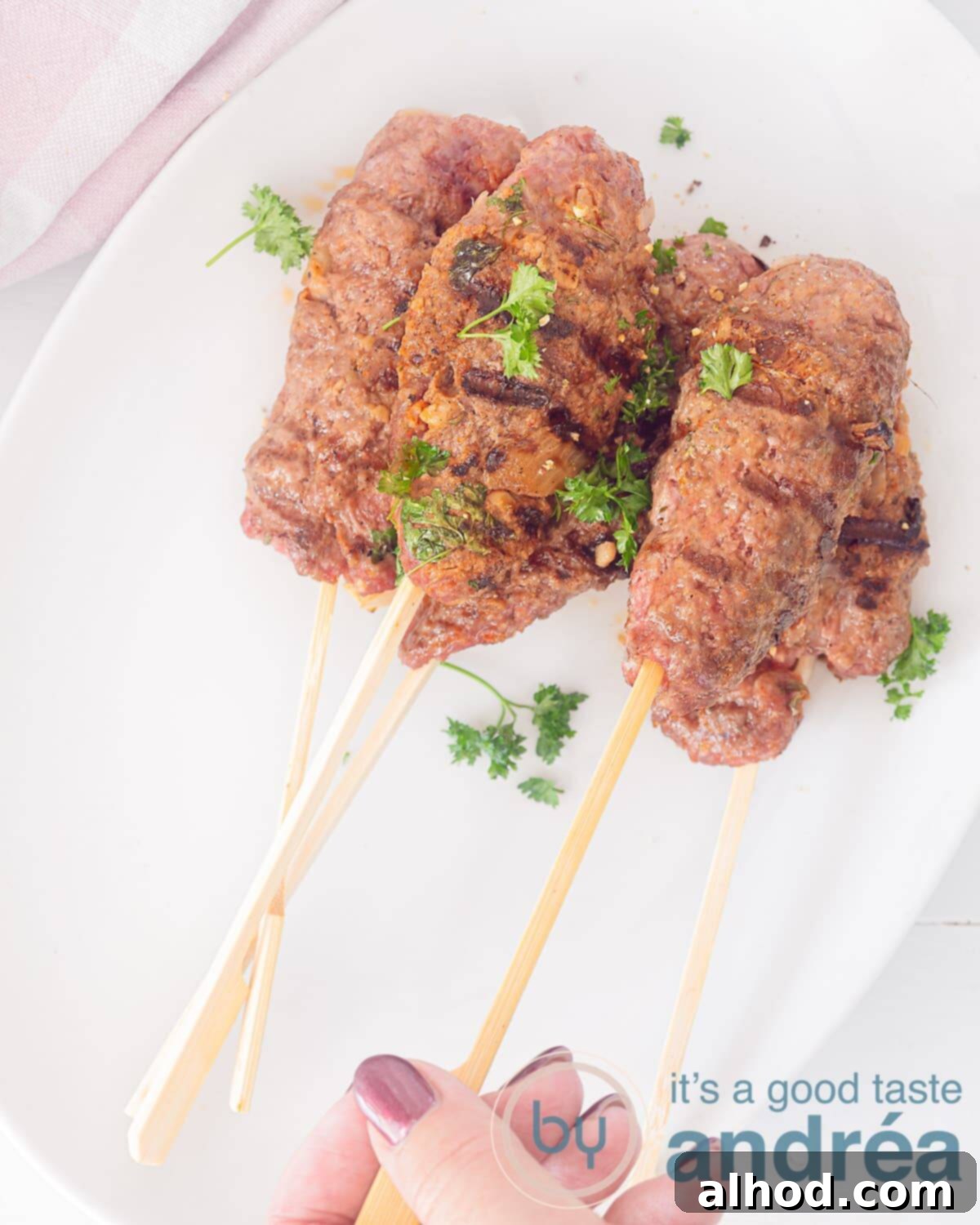 Homemade Moroccan Spiced Köfte Skewers 3 A high-angle photograph showing several köfte kebab skewers arranged on a white dish, with a hand reaching in to take one skewer.