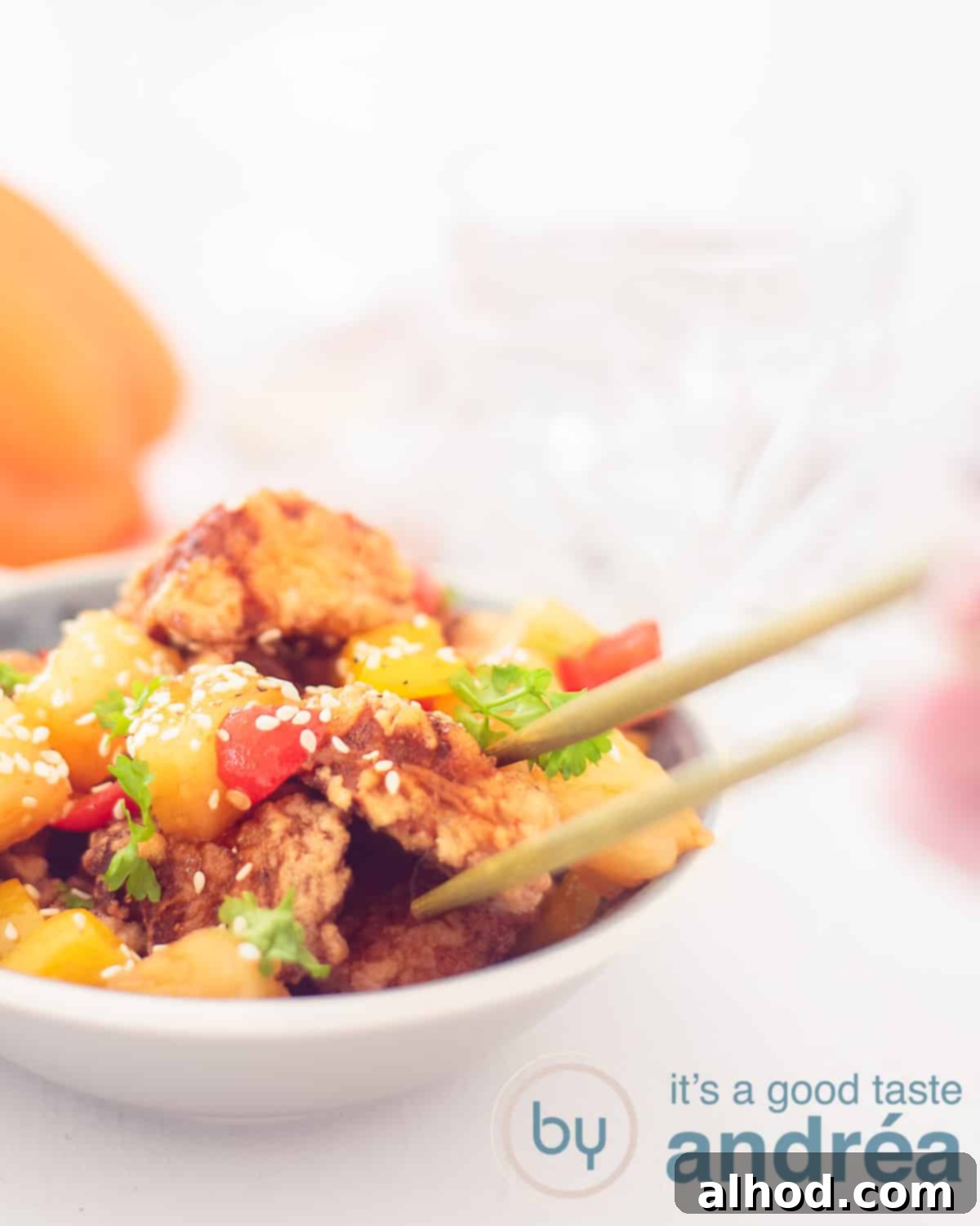 Sweet and Sour Pork 5 A height photo with a gray dish with Koe Loe Yuk in sweet and sour sauce on the left, garnished with sesame seeds. Two chopsticks grab a piece of meat from the dish. A clean white background with a yellow bell pepper on the right side.