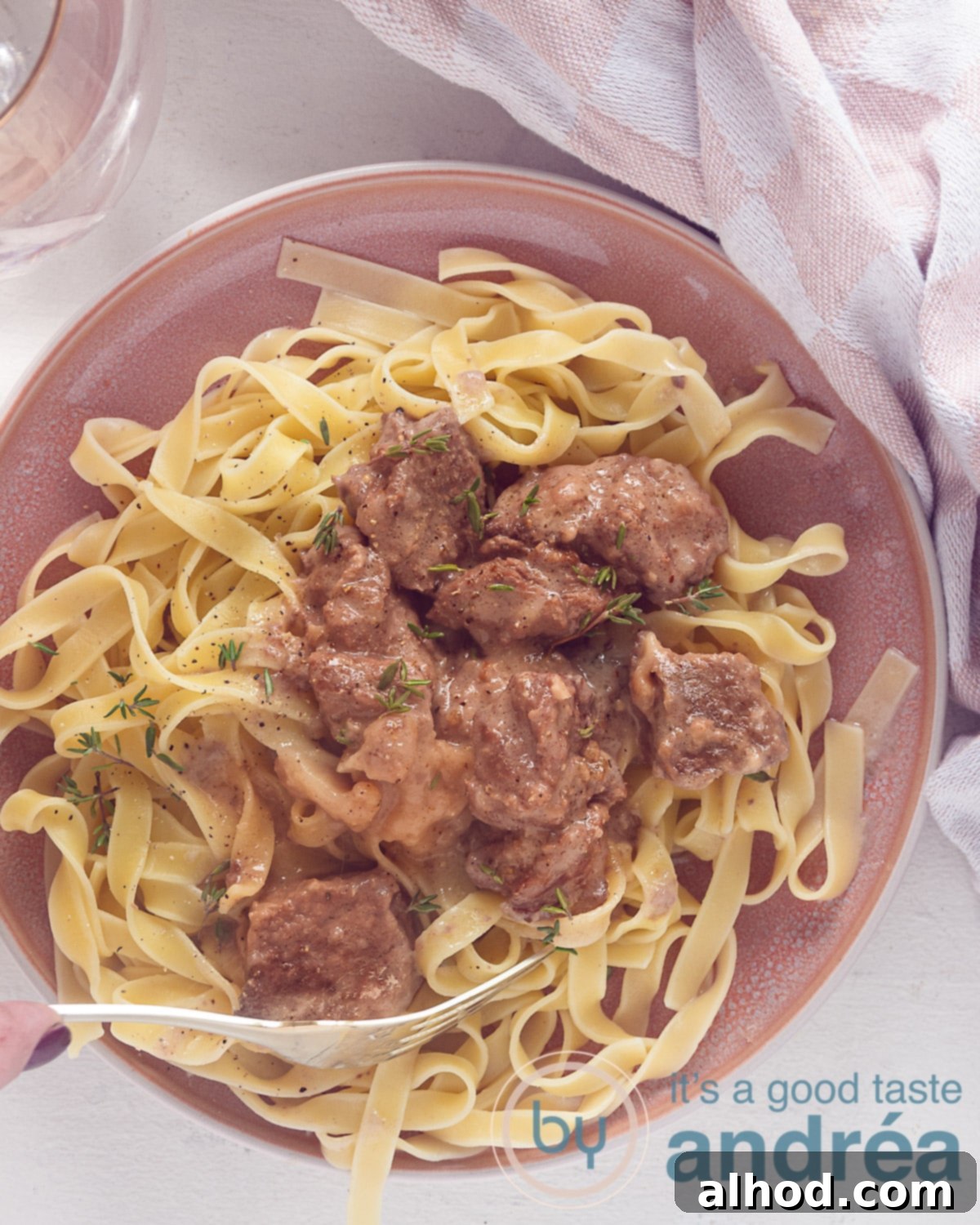 Slow Cooker Beef and Noodles 2 A photo from above of a pink plate with pasta with beef in a creamy sauce. A white background. At the top right a pink and white tea towel is draped.