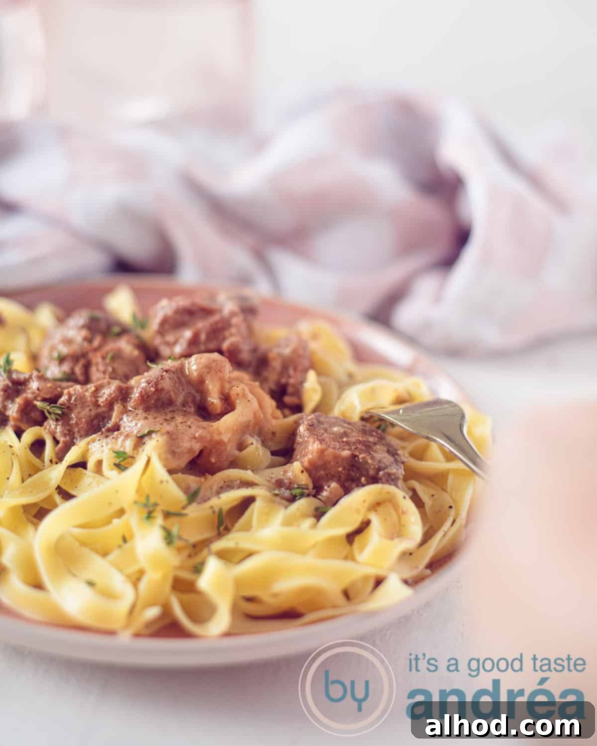 Slow Cooker Beef and Noodles 5 A pink plate of tagliatelle with a creamy beef sauce. A white background. A pink white tea towel in the background.