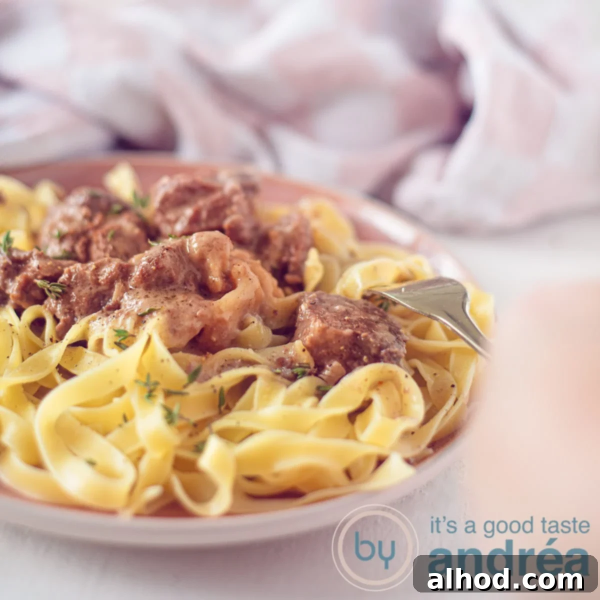 Slow Cooker Beef and Noodles 6 A square photo with a pink plate with pasta and beef in a creamy sauce. A hand pierces the flesh. A pink white tea towel in the background.