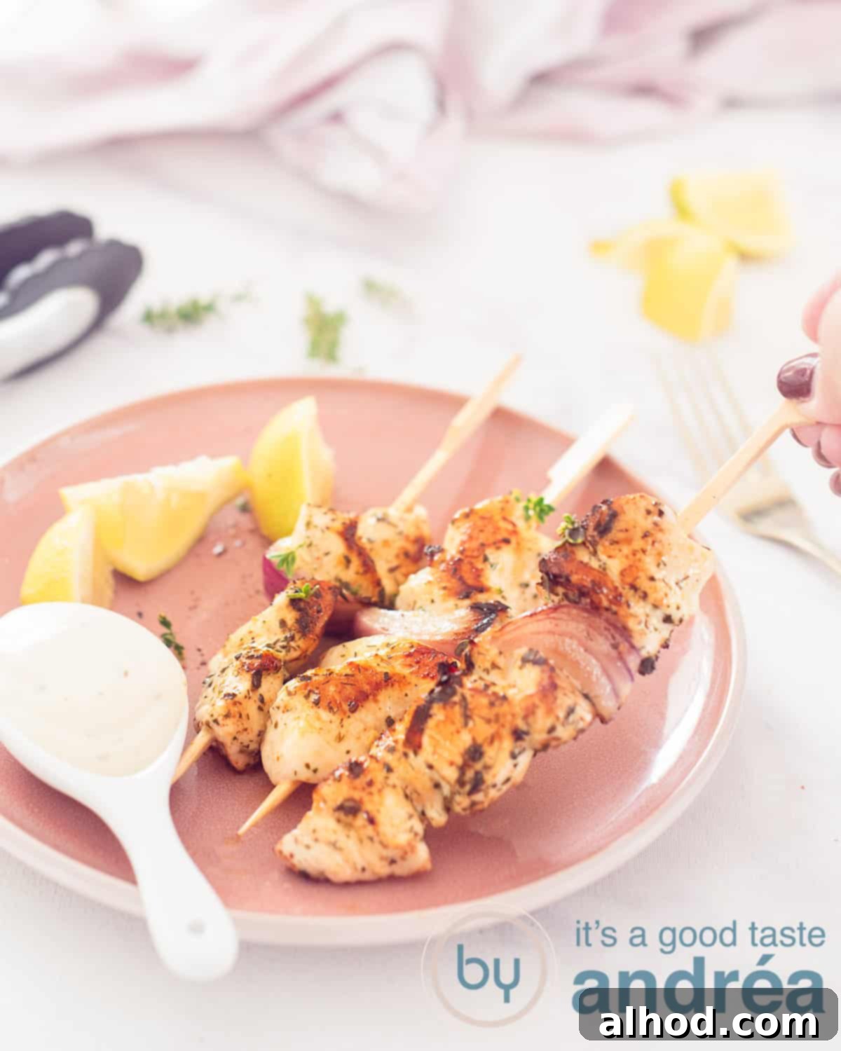 A pink plate with three Greek chicken skewers, lemon wedges and a white spoon with garlic sauce. A hand takes a stick. A white background. In the background more lemon and a pink white tea towel.