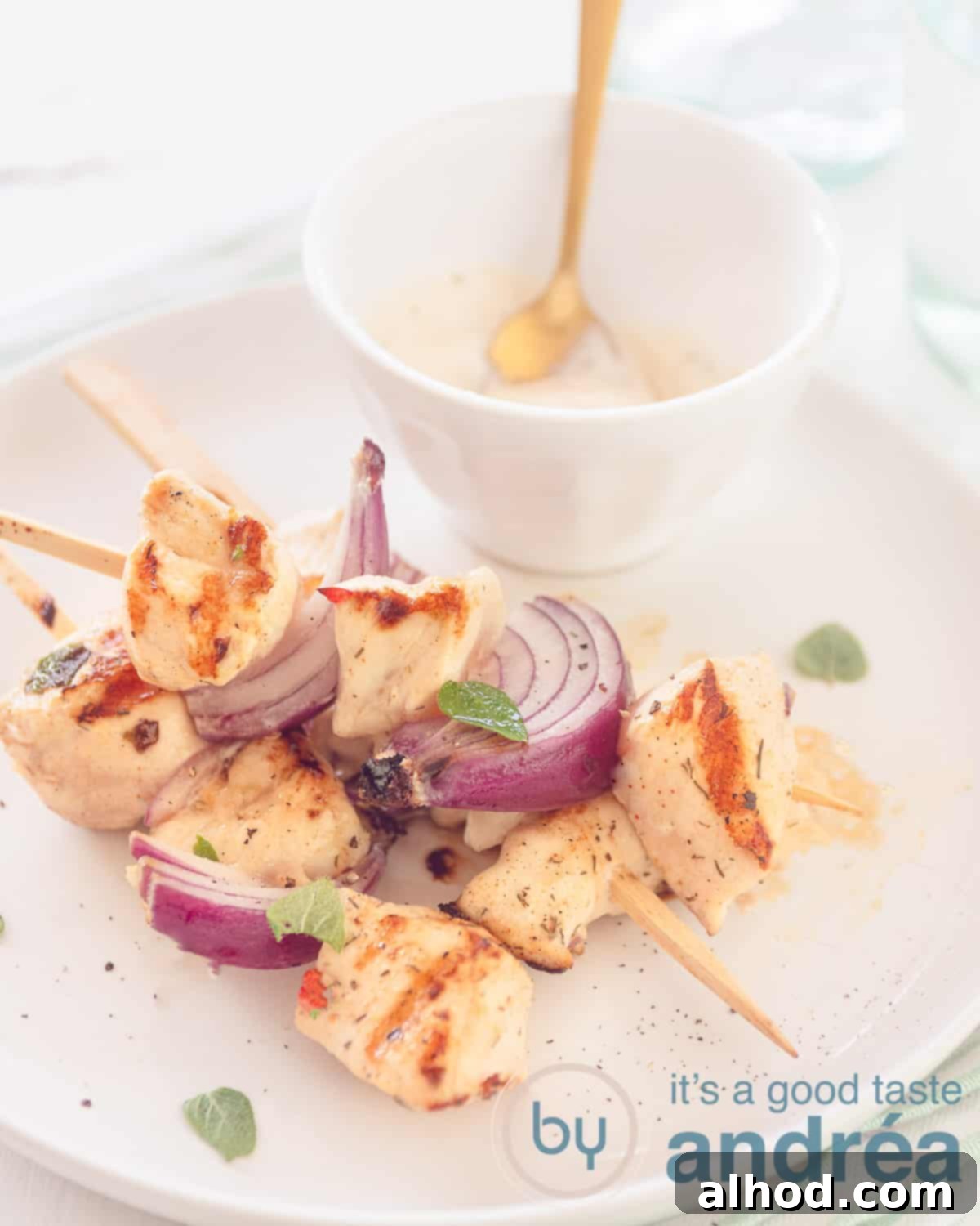 Flavorful Chicken Souvlaki Skewers 5 An elevation photo with a white plate on a white background with a bowl of tzatziki. On the plate three chicken skewers with chicken alternated with red onion.