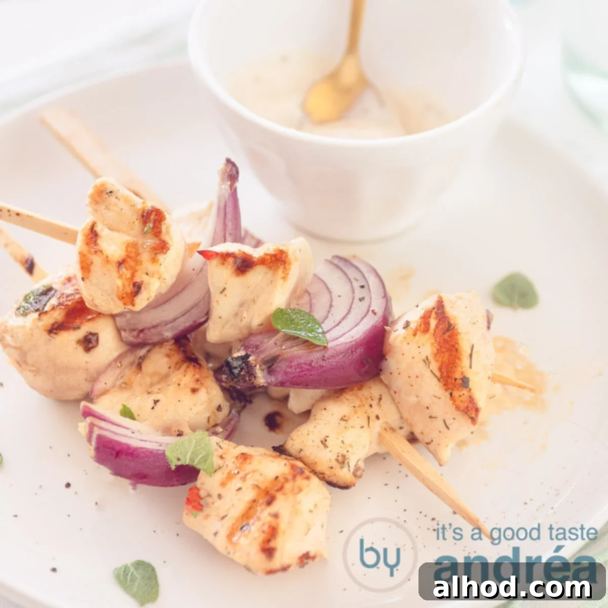 Flavorful Chicken Souvlaki Skewers 6 A white plate with chicken skewers with red onion. A bowl of tzatziki. A square photo.