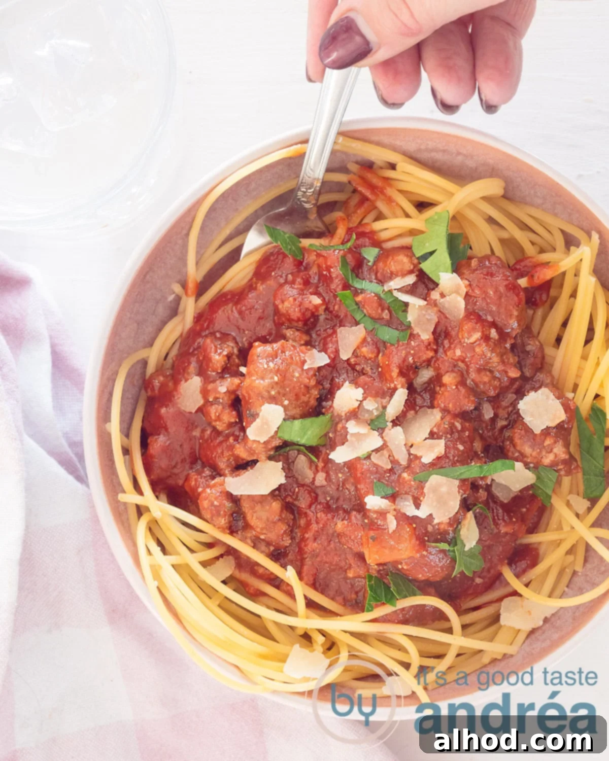 Ragù alla Bolognese 2 A photo from above of a bowl of spaghetti topped with homemade bolognaise sauce with Parmesan cheese and parsley. A hand holds a spoon. A white background, at the bottom left a white pink tea towel.