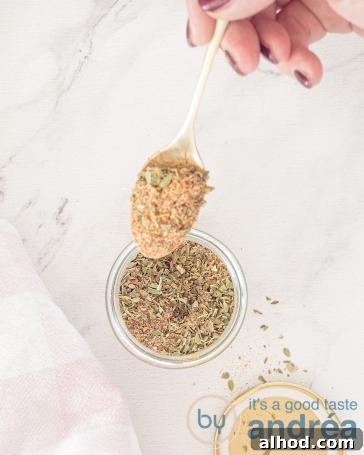 Authentic Homemade Gyros Seasoning 2 A photo from above with a glass jar with gyros spices. A golden spoon scoops spices from the glass. A white background. The lid is in front.