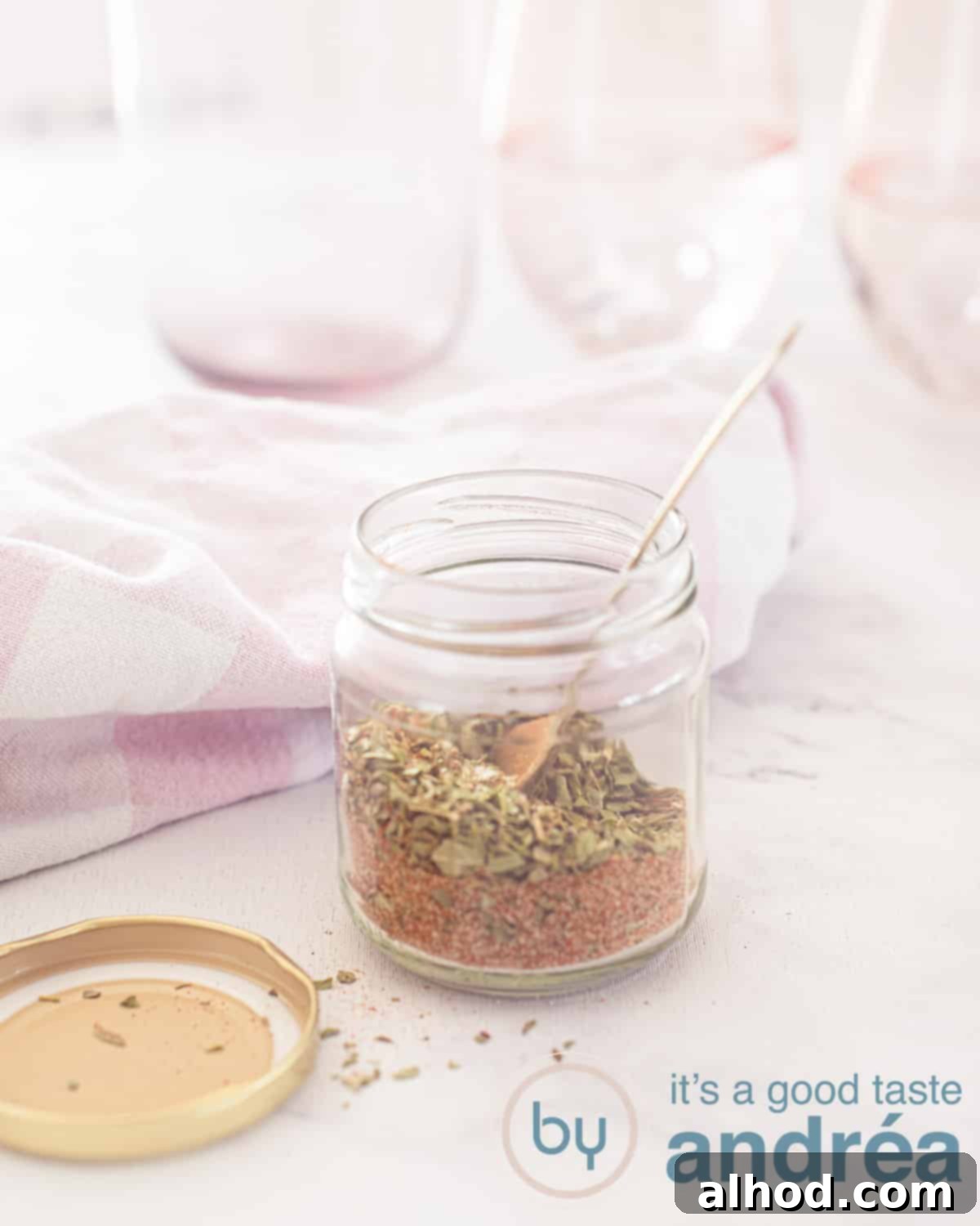 DIY Gyros Seasoning Blend 6 An elevation photo with a white background. In the middle a glass jar with Gyros spices, a golden spoon in the lid next to it. In the background pink glasses and a white pink tea towel.