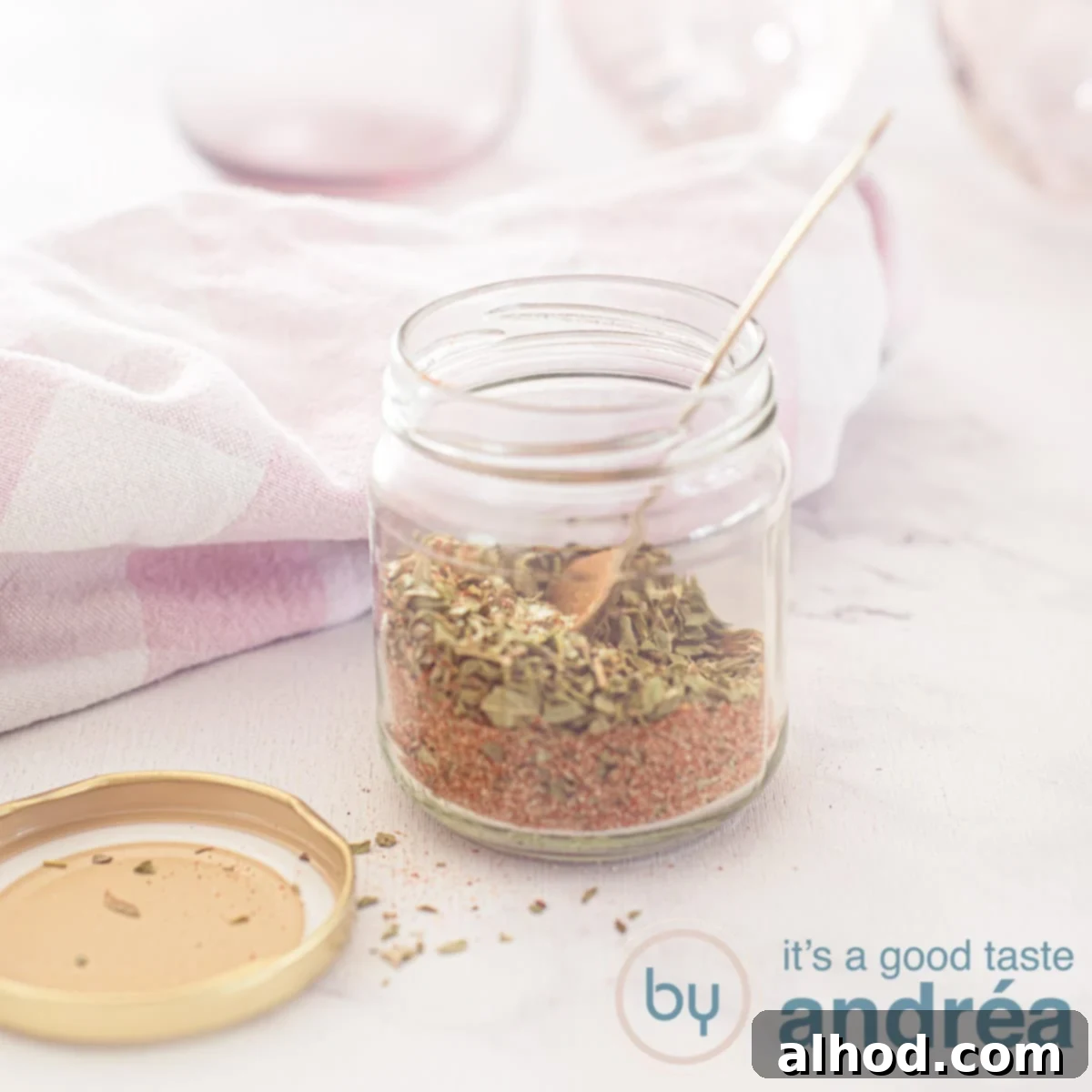 Authentic Homemade Gyros Seasoning 7 A square photo with a glass jar filled with gyros spice mix in the middle. A golden spoon is in it. The lid is next to it. A pink white tea towel and pink glasses in the background.
