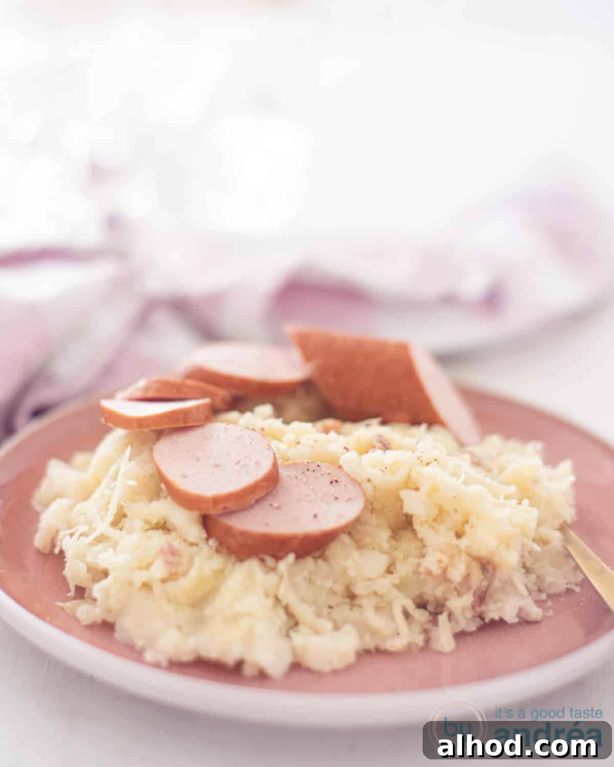 Hearty Dutch Sauerkraut Potato Mash 2 A photo of a pink plate with a scoop of sauerkraut mash, bacon and slices of smoked sausage on it. In the background a pink and white tea towel.