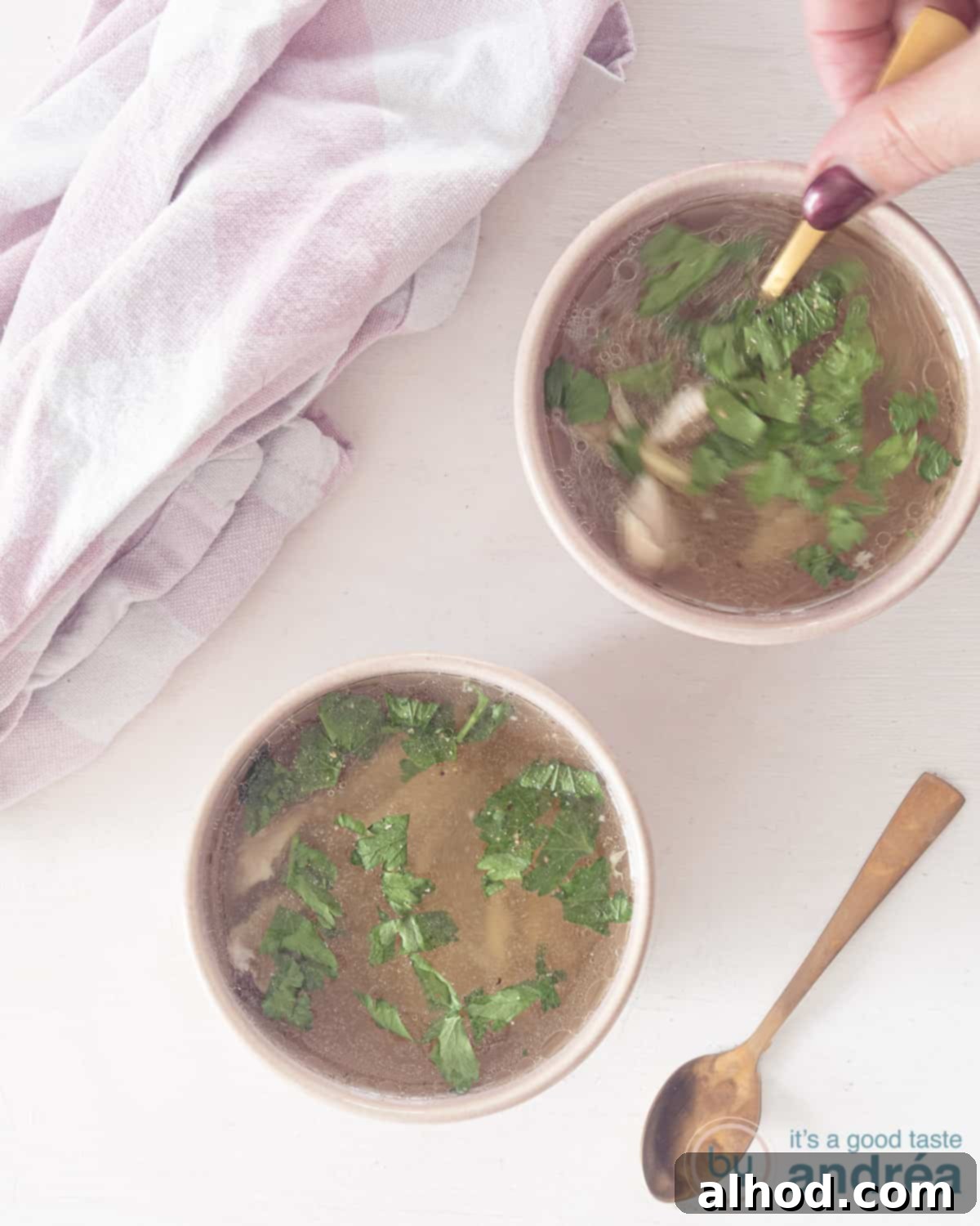 A photo from above of two bowls filled with fresh chicken soup made with chicken leg. A spoon scoops out a bite. A white bowl. Top left a pink and white tea towel.